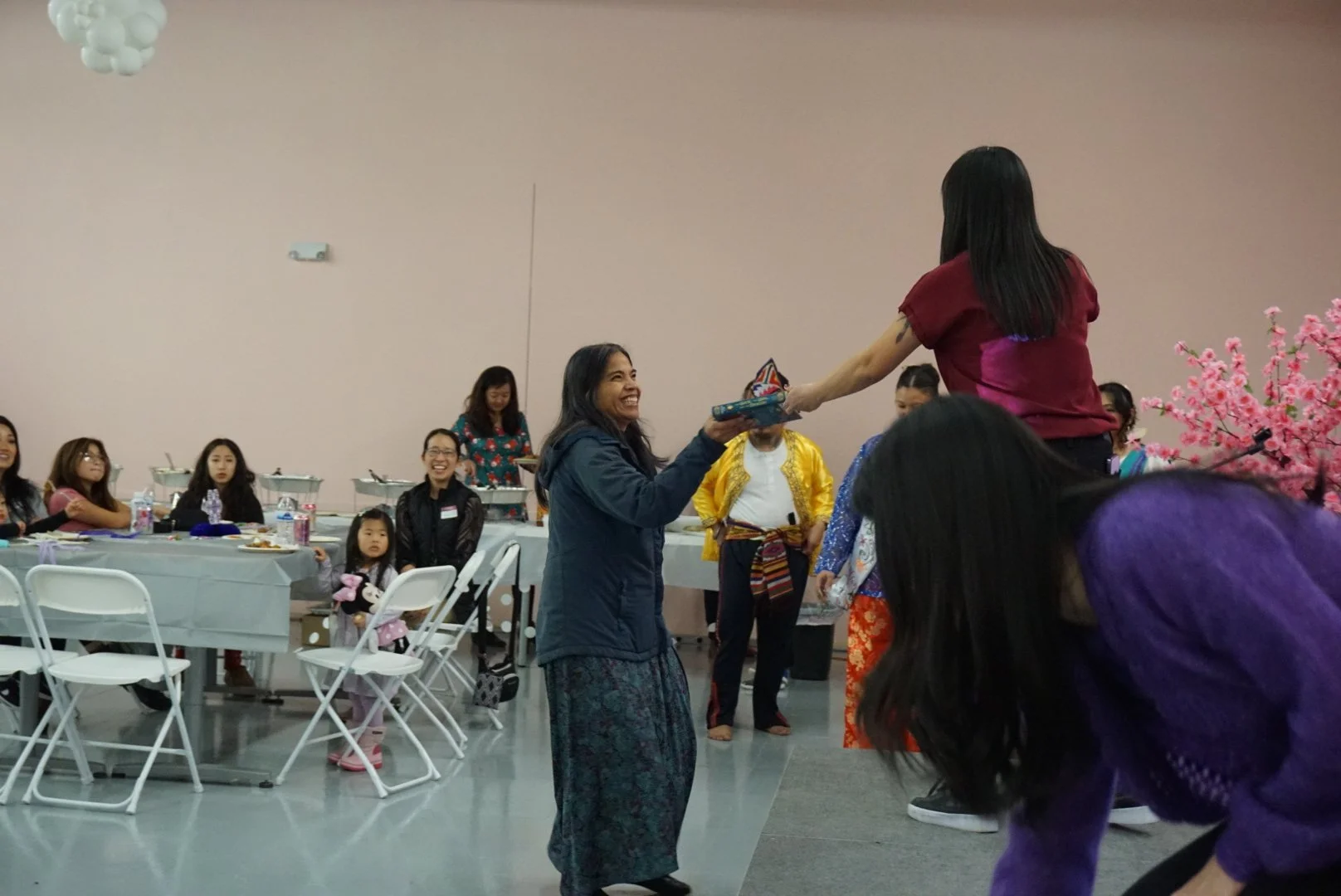  Family receiving raffle prize from Joanne Liu on stage in light pink room with folding chairs, tables, and community  