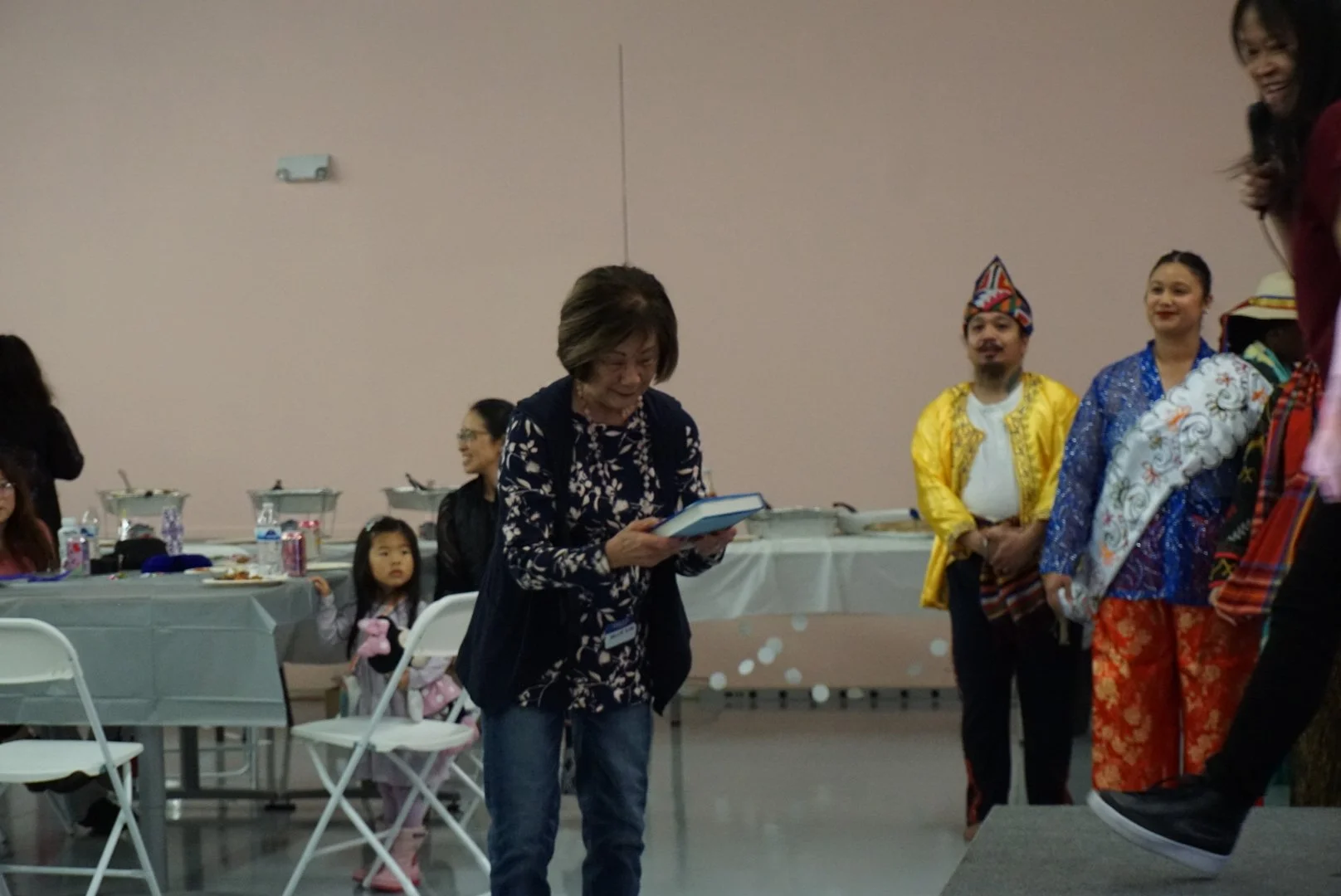  Family receiving raffle prize from Joanne Liu on stage in light pink room with folding chairs, tables, and community  