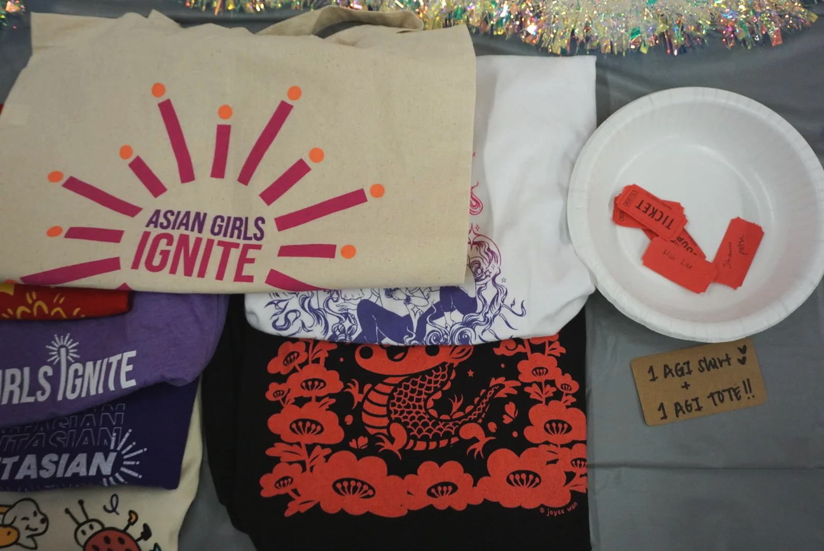  AGI shirts and tote bag folded on left side next to paper bowl with tickets for raffle on gray table with tinsel decor 