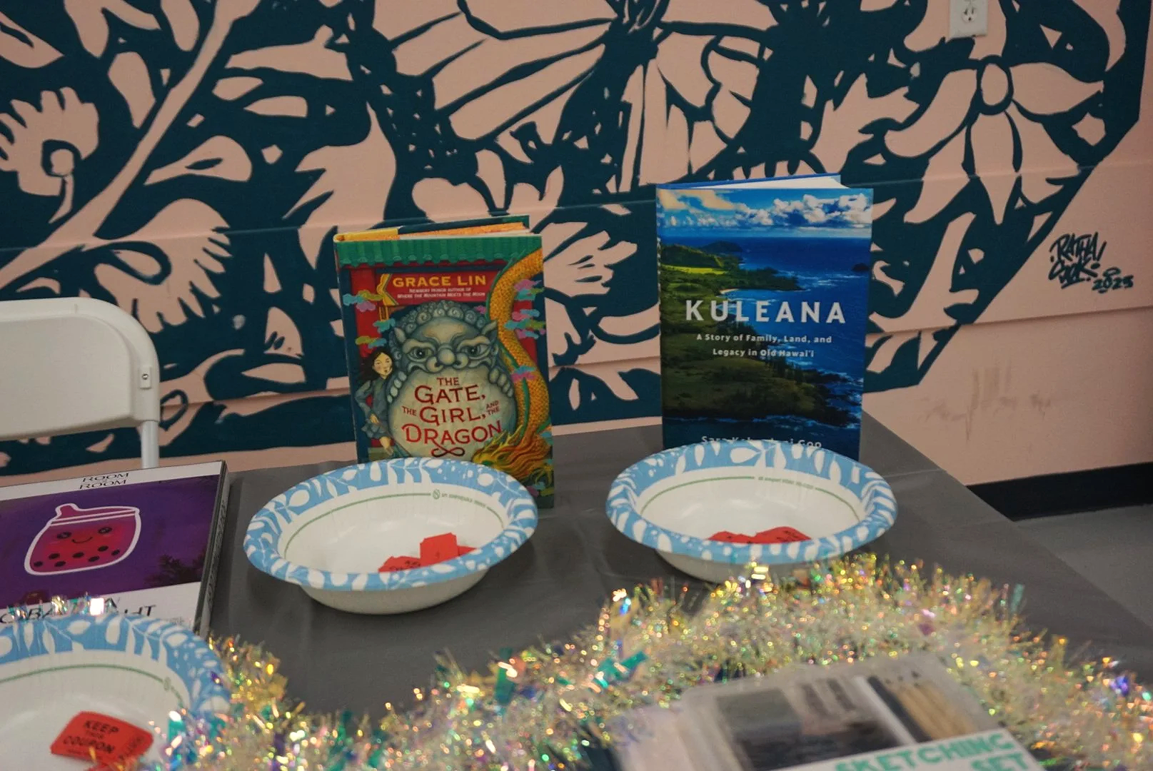  Grace Lin’s “The Gate, the Girl, and the Dragon” book standing behind paper bowl with tickets next to Sara Kehaulani Goo’s “Kuleana” book standing behind paper bowl with tickets on raffle table 