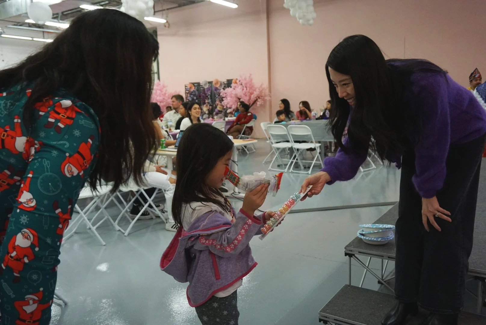  Family receiving raffle prize from Mehgan Yen on stage in light pink room with folding chairs, tables, and community  