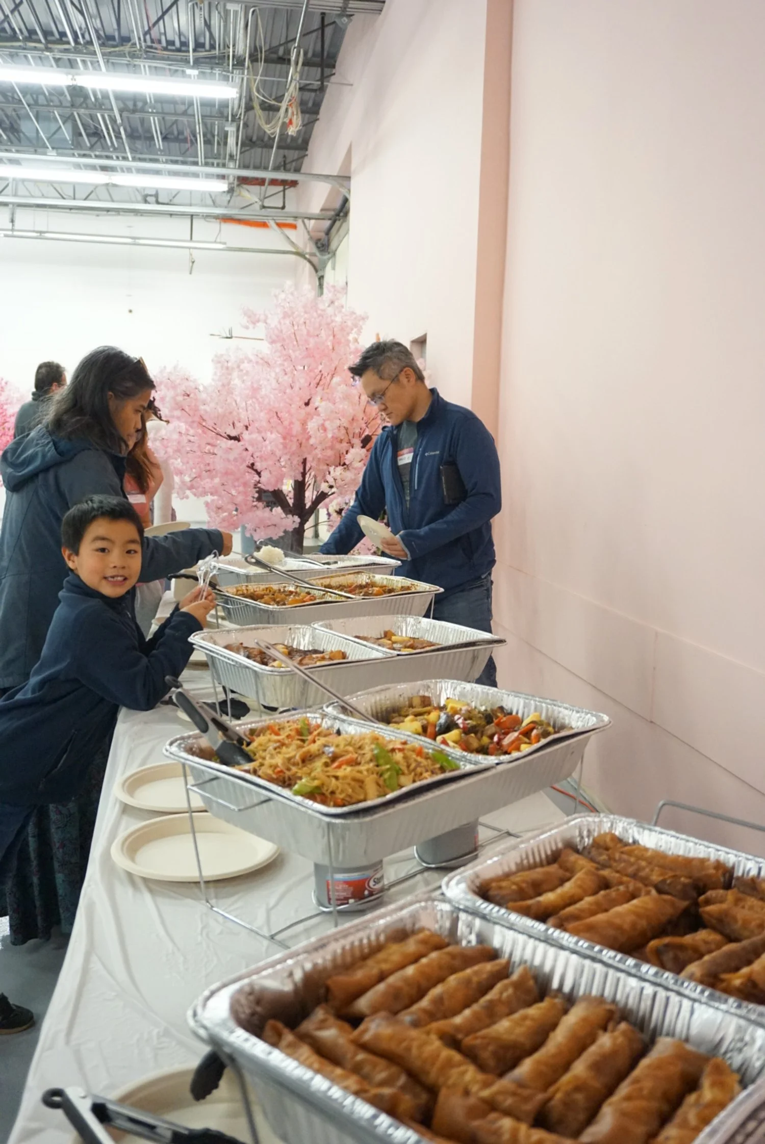  Families serving food in pink room with faux cherry blossom tree behind table with tin trays of food  