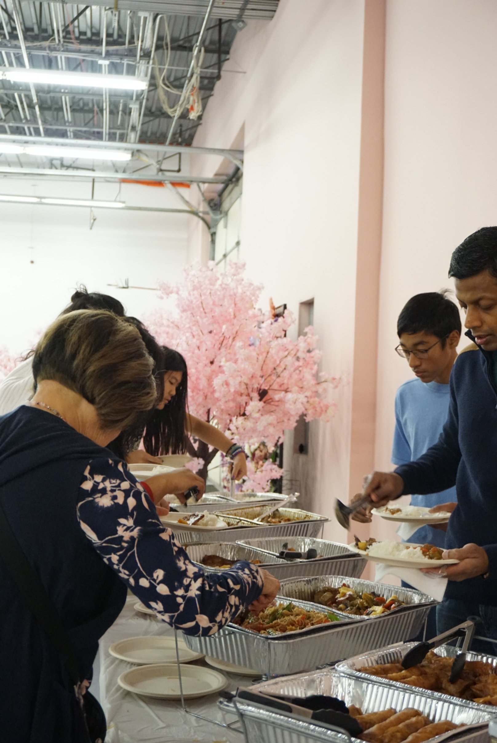  Families serving food in pink room with faux cherry blossom tree behind table with tin trays of food  