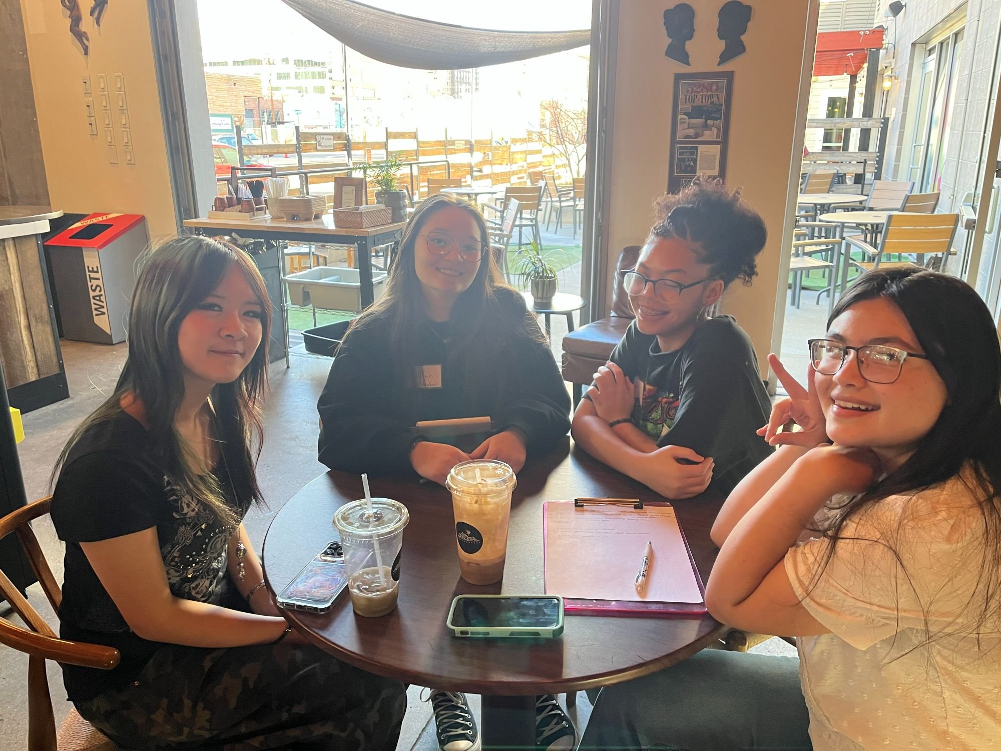  Four students sit together at a table and pose for a picture with drinks, a clipboard, and phones on the table. 