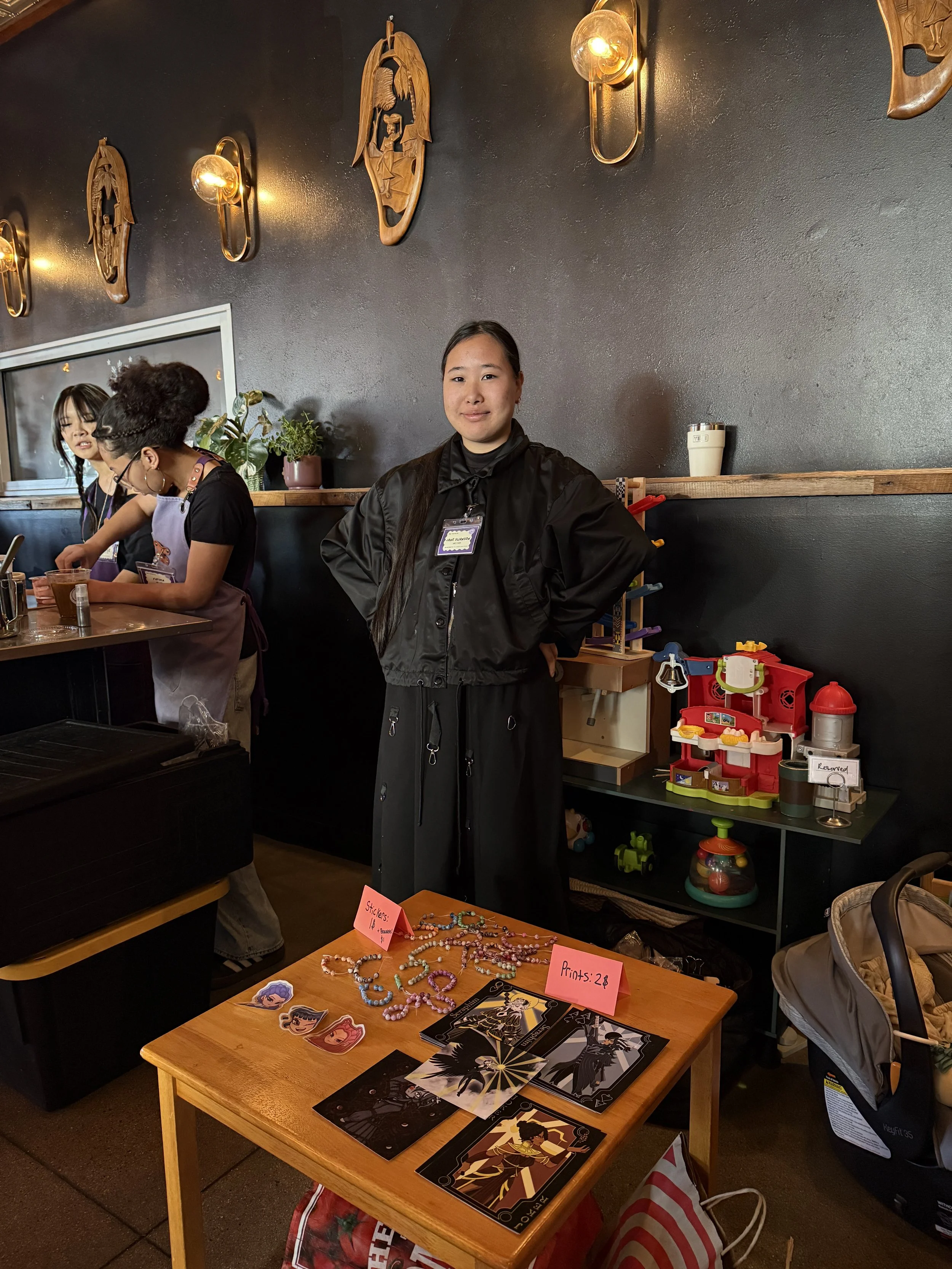  Jane S. stands at table with their artwork at Coffee Sarap. 