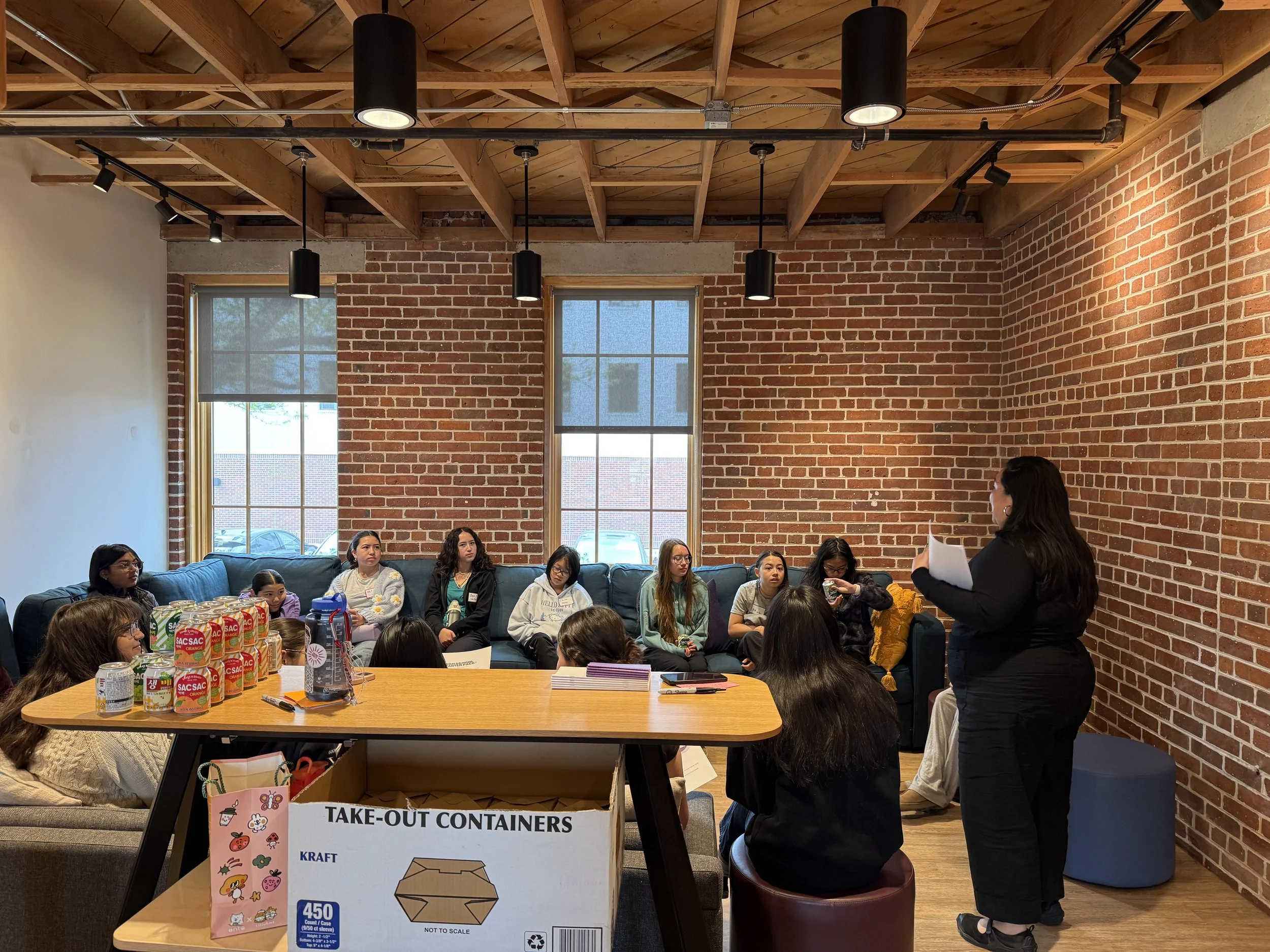  Jihee Yoon speaks to AGI middle school students seated on blue couch at High School Starter Guide Event in brick wall room and tall windows behind couch 