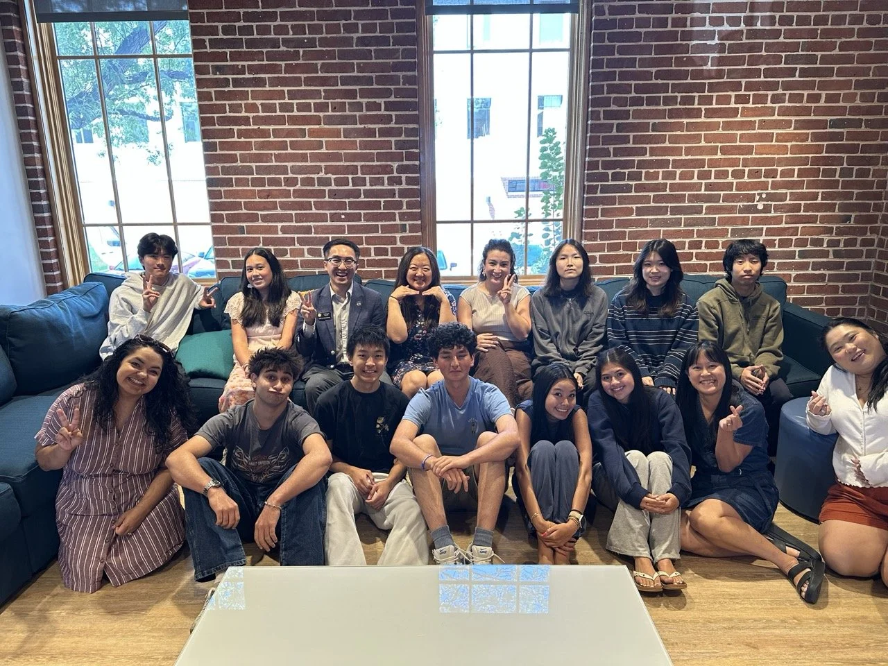 Group photo of AANHPI Youth Leadership & Advocacy Program students, staff, and guest speakers seated together in a brick-walled room, several making peace signs