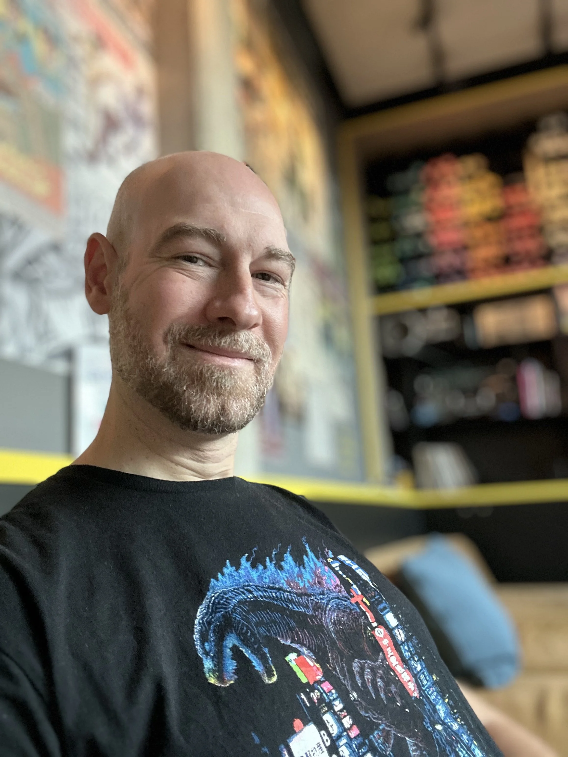 Ein lächelnder Mann names Daniel mit Bart und Glatze posiert in einem Raum mit bunten Postern und Büchern im Hintergrund.