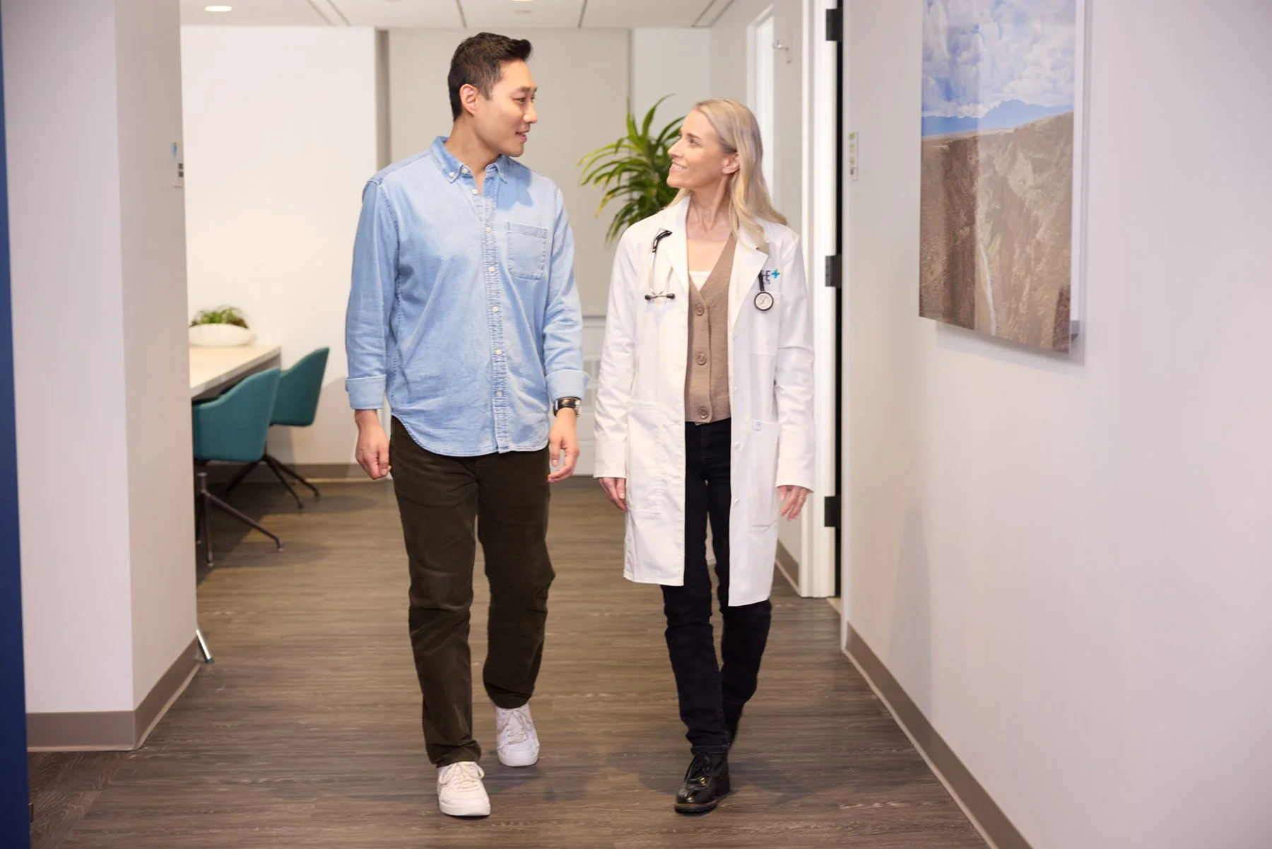 A healthcare professional, dressed in a white coat with a stethoscope, walks and talks with a young man in casual clothing in a medical facility hallway. They are smiling at each other.