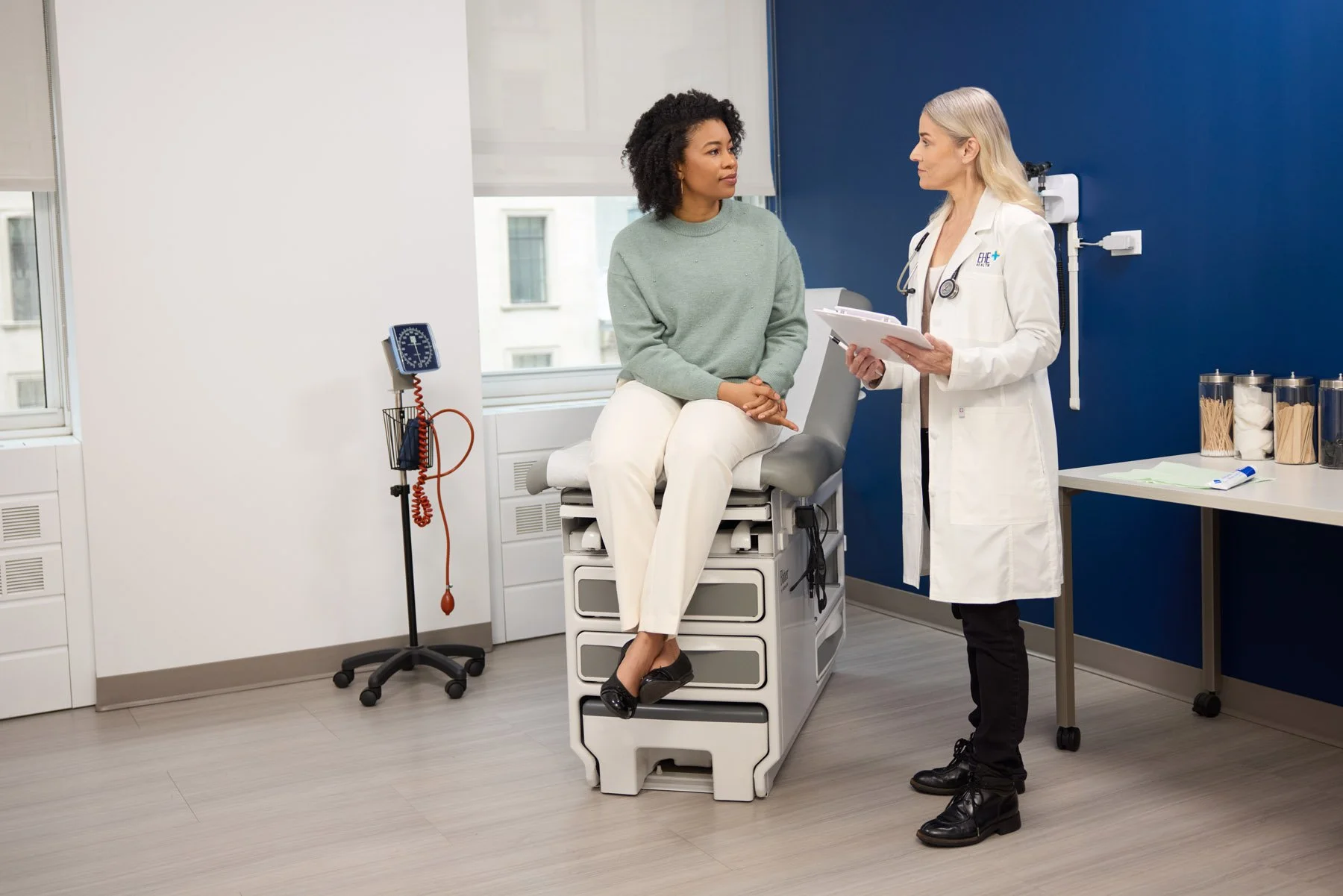 A patient sitting on an examination table in a medical clinic, talking with a female doctor who is standing beside her with a clipboard. The clinic room has white and blue walls, medical equipment, and supplies in the background.