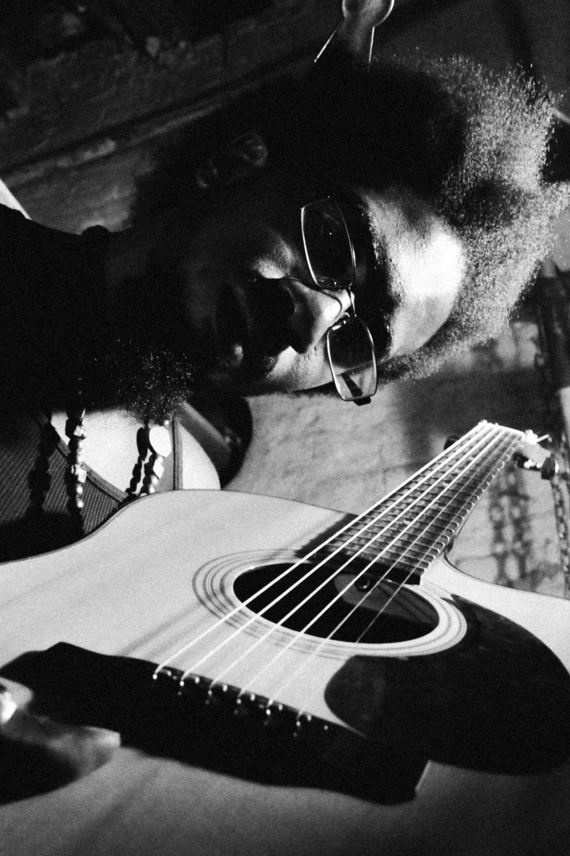 Black and white photo of a person with glasses playing an acoustic guitar, wearing a beaded necklace.