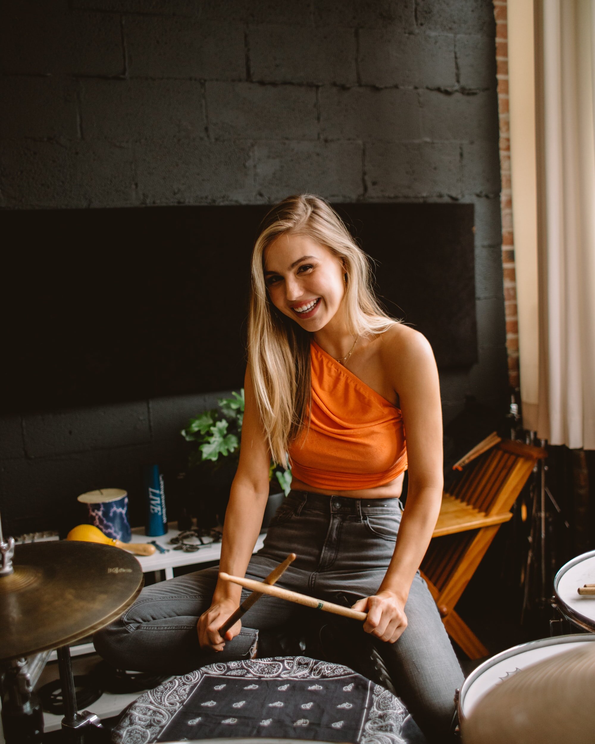 Woman smiling and holding drumsticks, sitting at a drum set indoors, wearing an orange top.