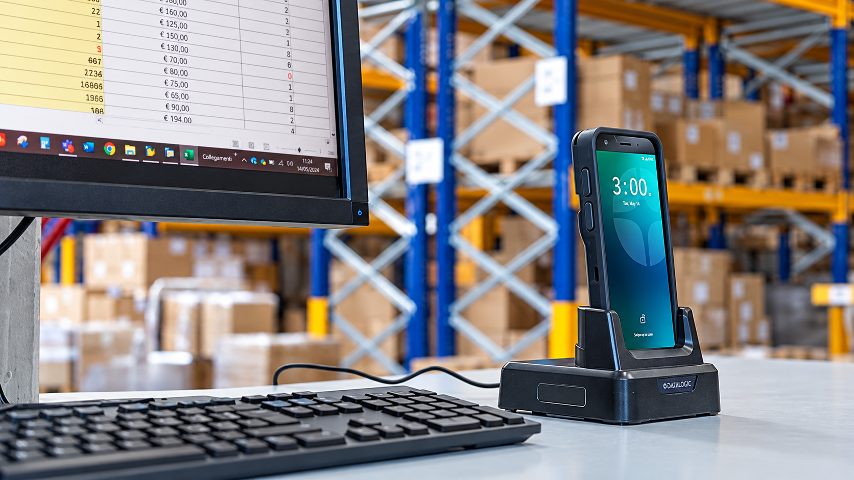 Datalogic Memor mobile computer docked at a warehouse workstation beside a keyboard and monitor, with storage racks and boxes in the background.