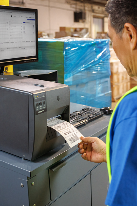 Warehouse worker pulling a printed barcode label from a Zebra ZT111 industrial label printer at a packing or shipping workstation.