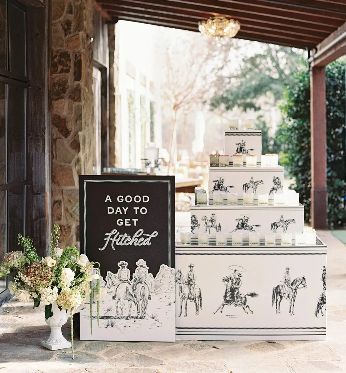 Decorative display of small jars with black and white horse illustrations, a bouquet of flowers, a black sign with white text that reads "A GOOD DAY TO GET HITCHED," and a tiered cake resembling a box with horse illustrations, set in a rustic outdoor setting with stone walls and greenery.