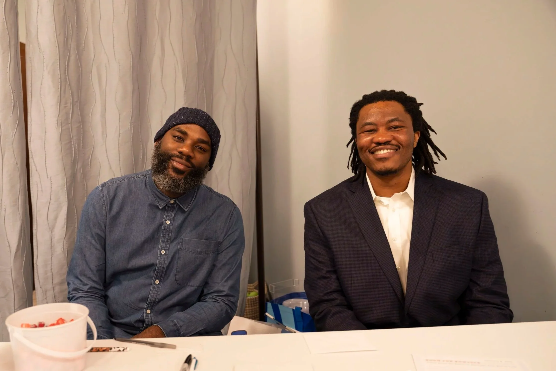 Two men sitting behind a table with various papers, pens, and a bucket of red items. One man wears a dark blazer and white shirt, smiling, and the other wears a denim shirt and beanie, also smiling. There are curtains and a plain wall behind them.
