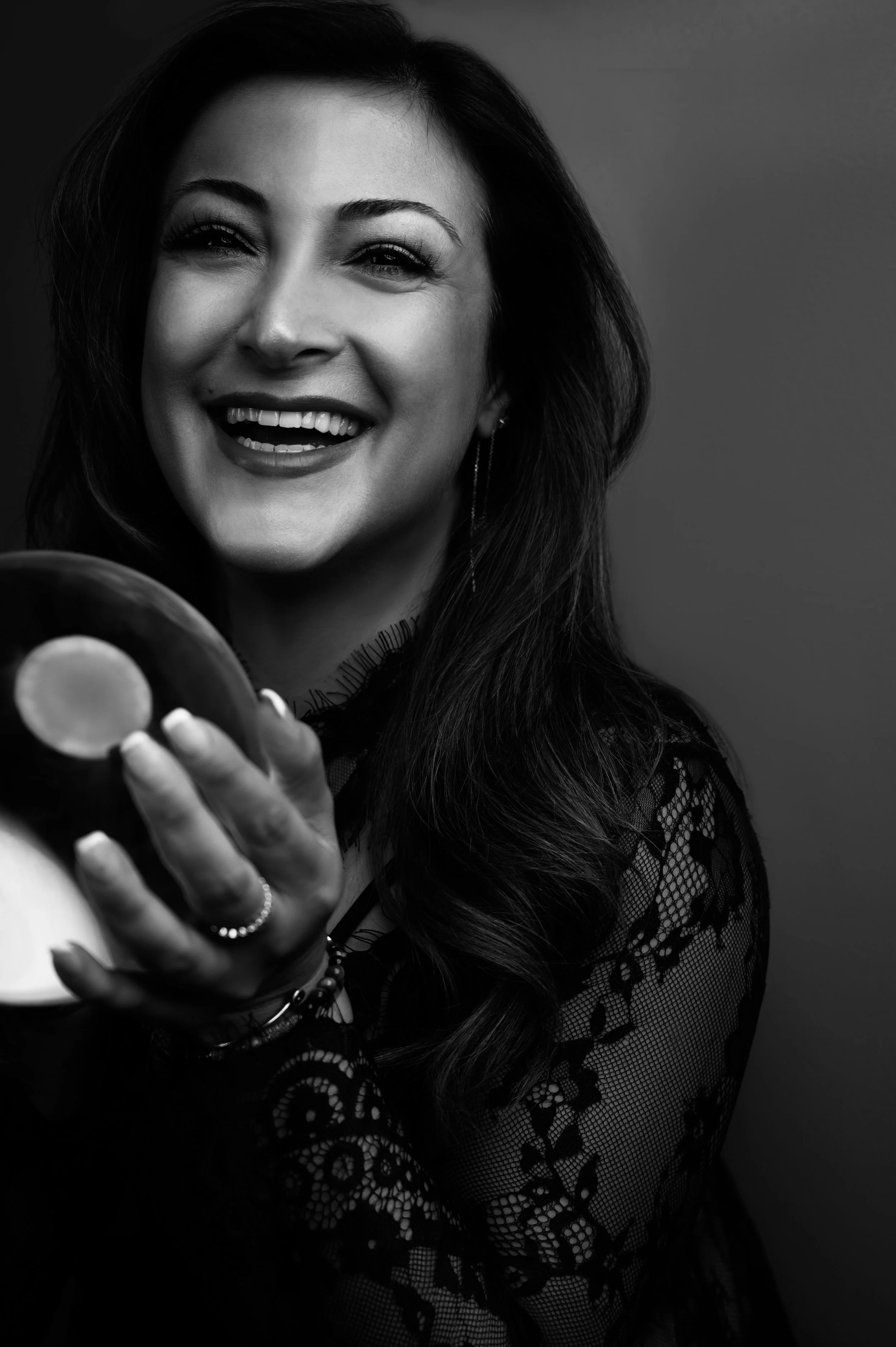 Black and white photo of a smiling woman with long, wavy hair holding a donut towards the camera, wearing a lace top and jewelry.