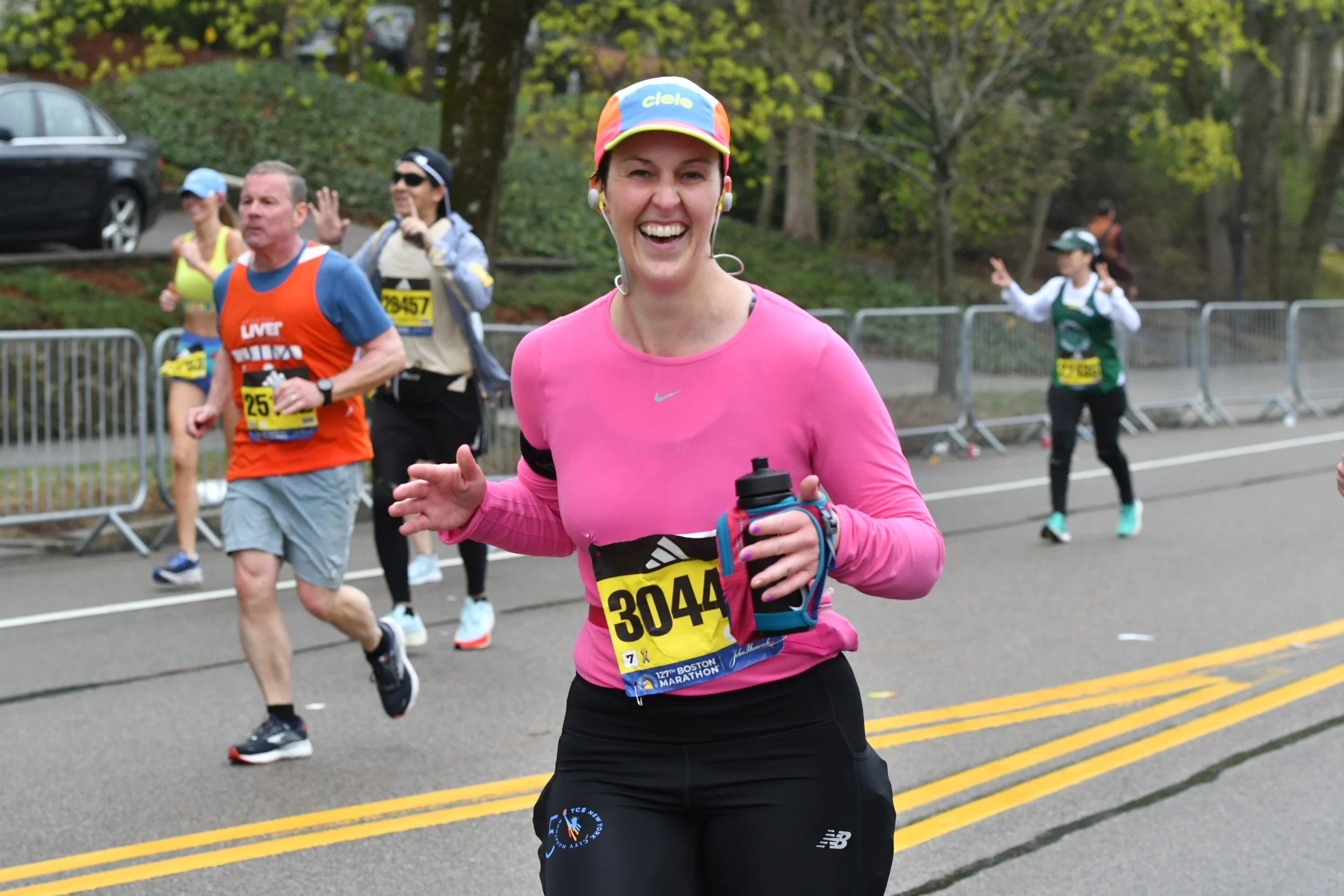 A woman smiling and running in a marathon with other runners behind her on a road lined with trees and barriers.