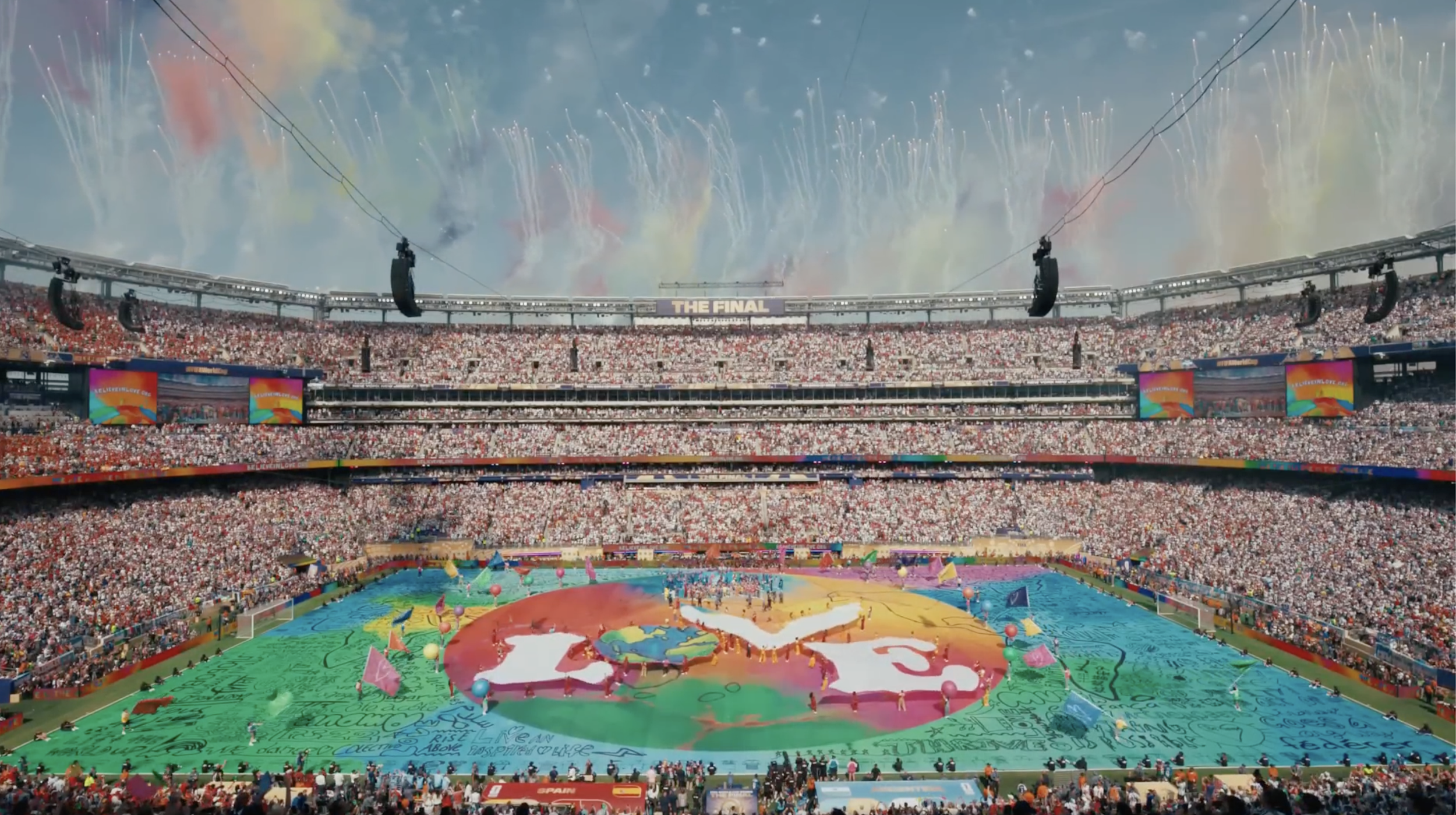 A large stadium filled with spectators for a sports event, with colorful fireworks in the sky and a large banner that reads 'The Final'. The field is decorated with a vibrant, artistic display that includes a world map and messages of unity, celebration, and peace.