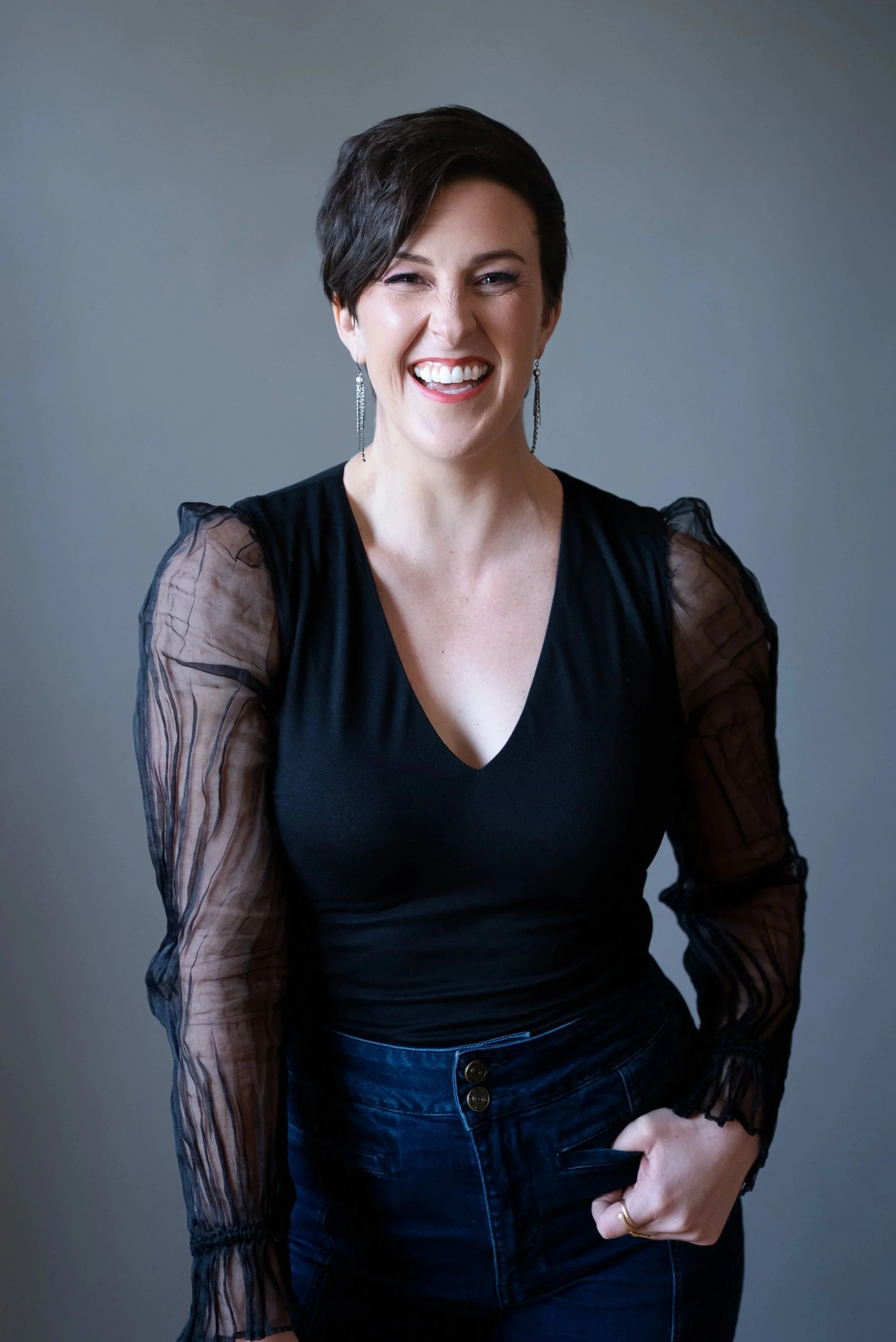 A woman with short dark hair smiling and wearing a black top with sheer, ruffled sleeves and earrings, standing in front of a plain gray background.