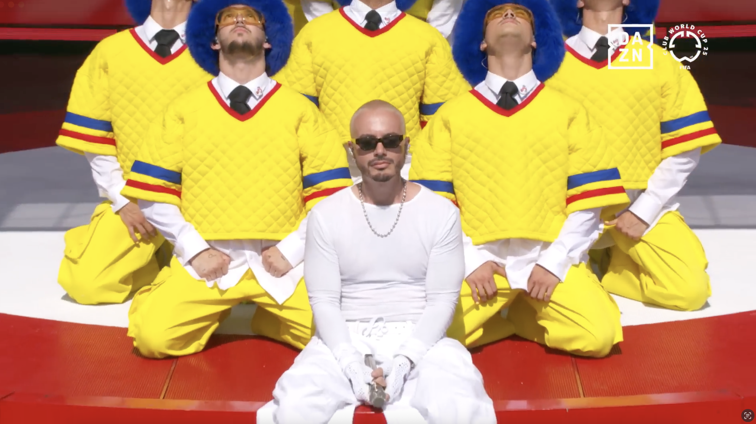 A group of six men dressed in yellow football uniforms with red and blue accents, kneeling on a red sports mat, with one man in white with sunglasses sitting in front of them. The scene appears to be at a sports event, with the NFL and Club World Cup logos visible.