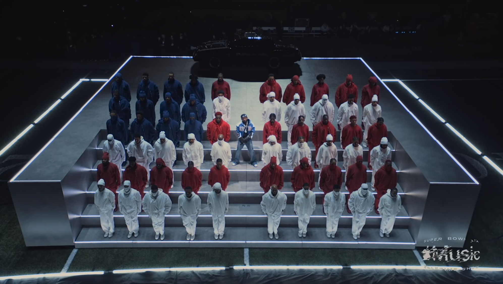 People dressed in white, red, and blue tracksuits standing on a raised, illuminated platform during a nighttime event, with a black vehicle and a crowd in the background.
