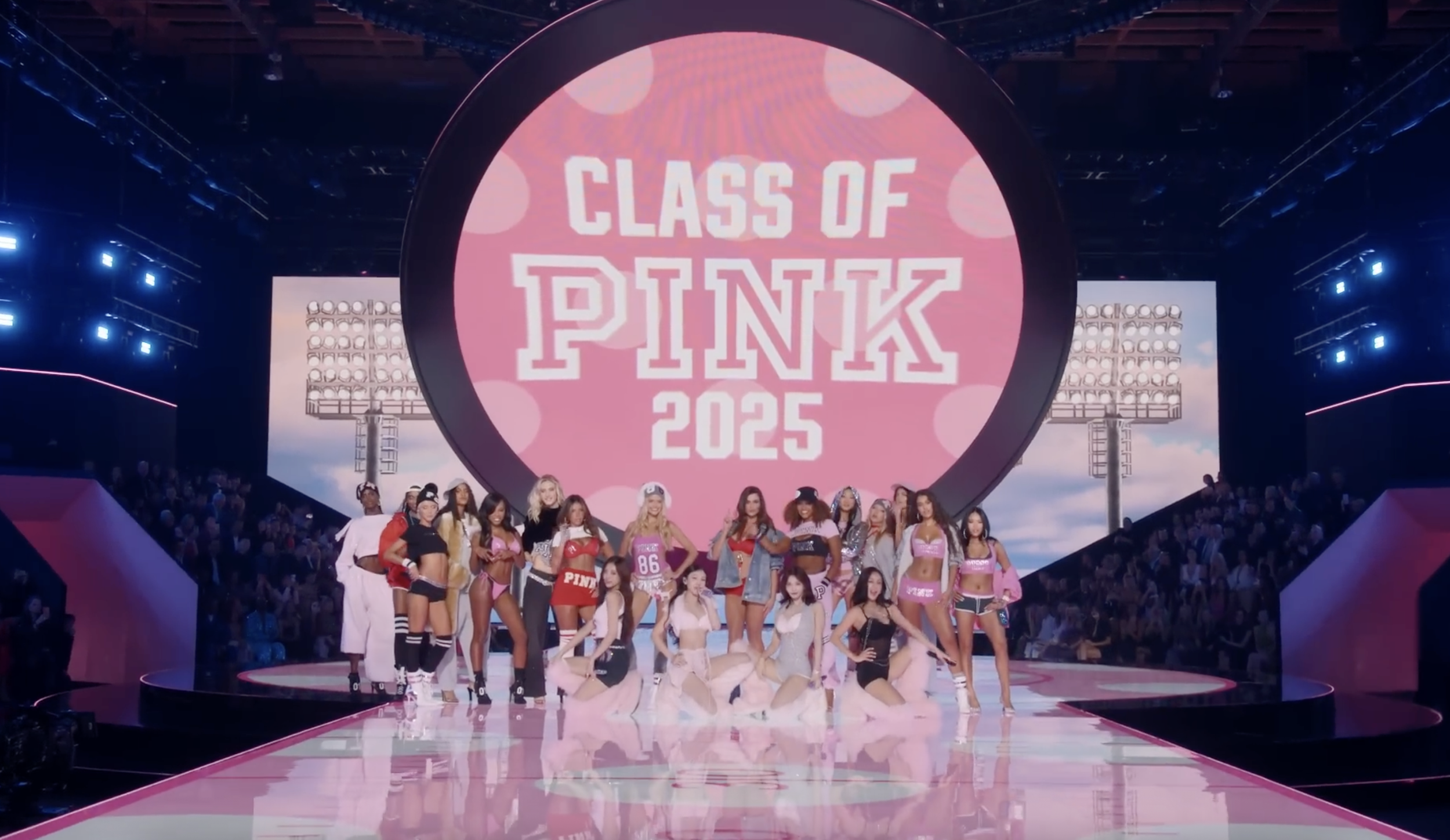 A group of young women and children in colorful athletic clothing on a runway during a fashion show called 'Class of Pink 2025', with a large pink circular display in the background.
