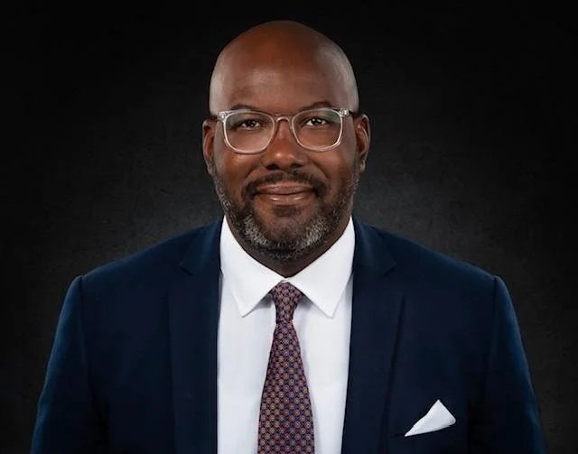 A portrait of a bald African American man with glasses, wearing a navy suit, white shirt, and patterned tie, against a dark background.