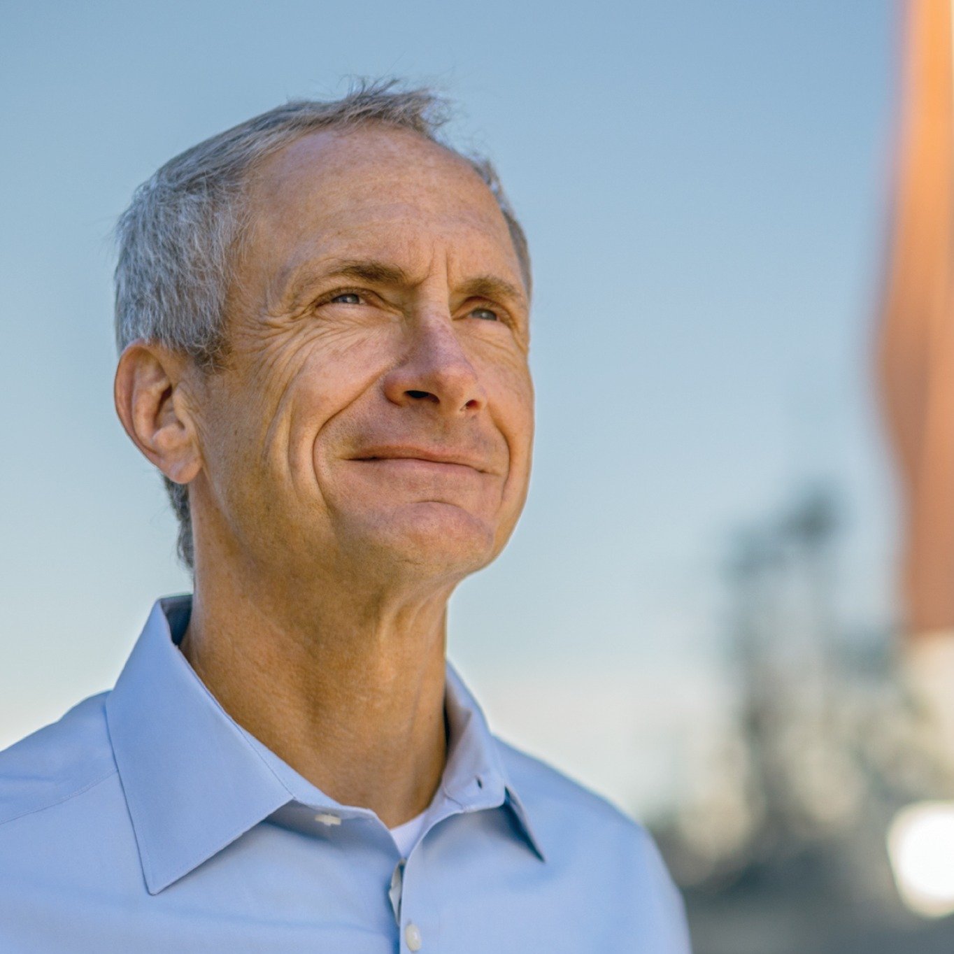 A middle-aged man with gray hair and blue eyes, smiling and looking towards the sky, wearing a light blue button-up shirt with blurred cityscape background.