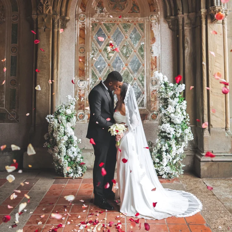 Newlyweds in a stone church doorway framed by white florals and falling petals
