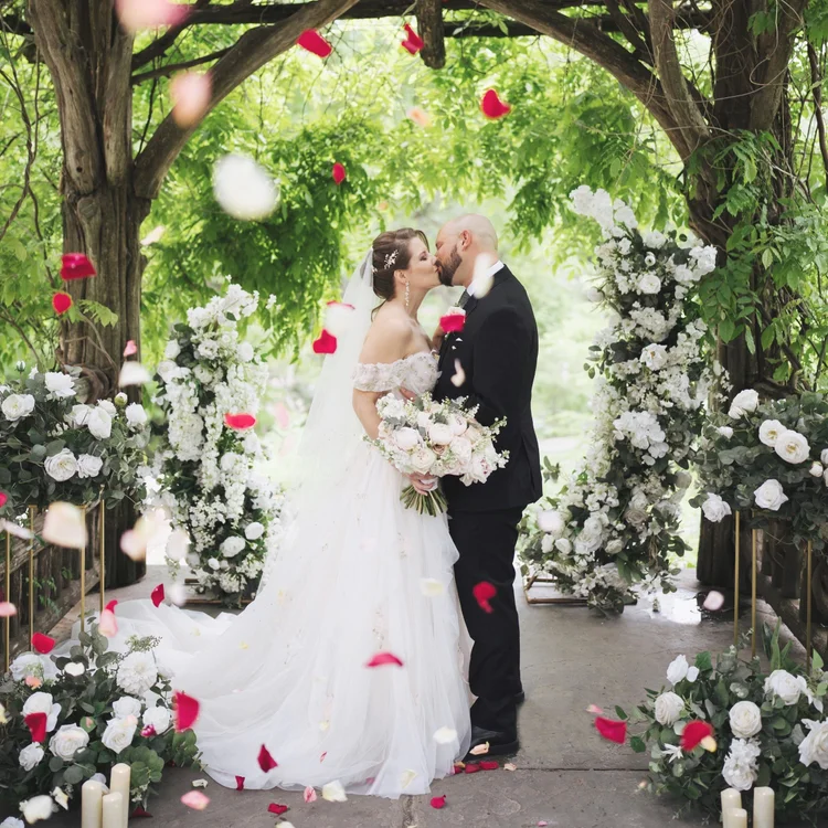 Couple kissing under a white and red floral arch as rose petals fall