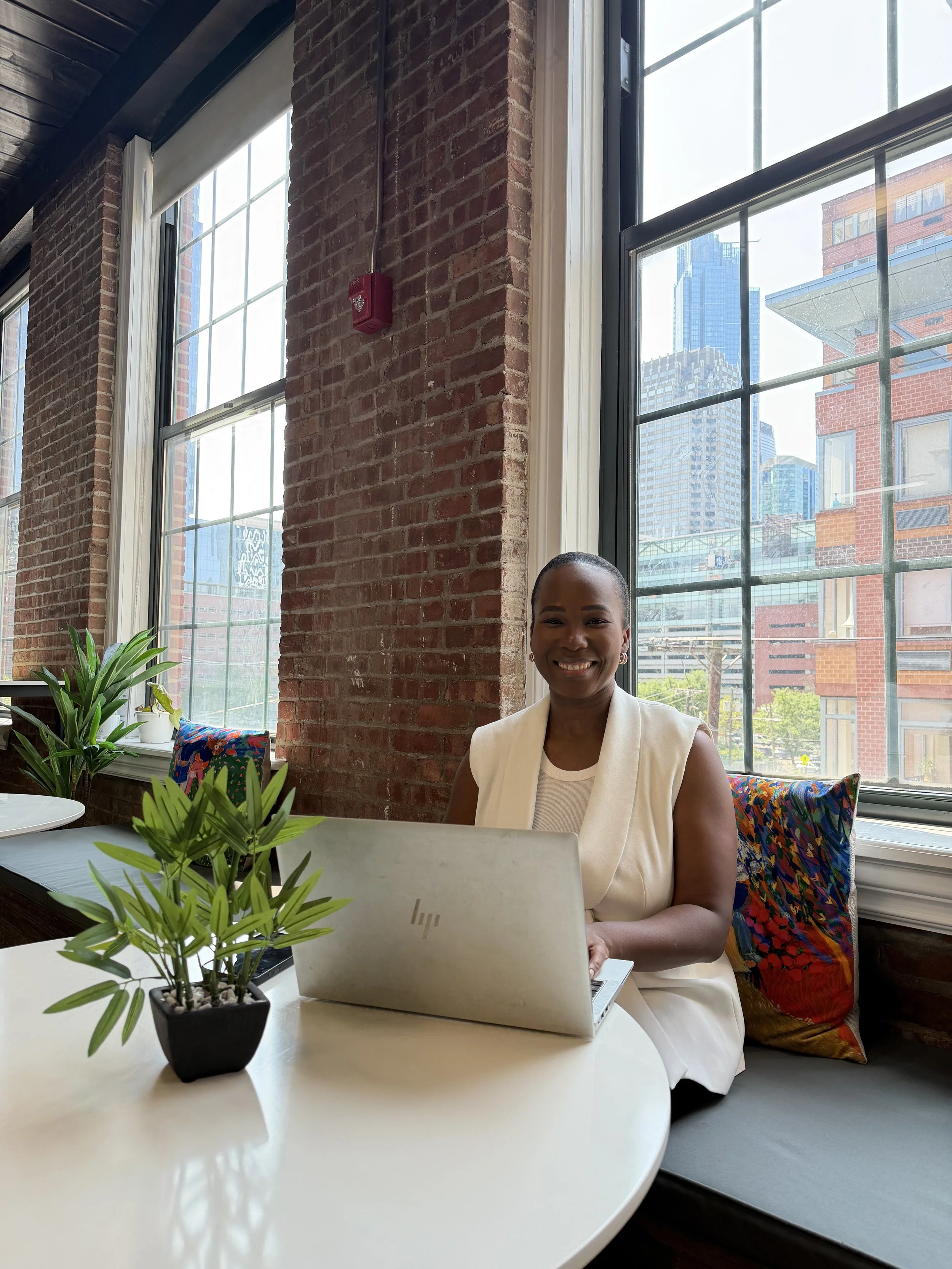 A smiling woman sitting at a white table with a potted plant, working on a laptop, in a bright room with large windows and a city view.