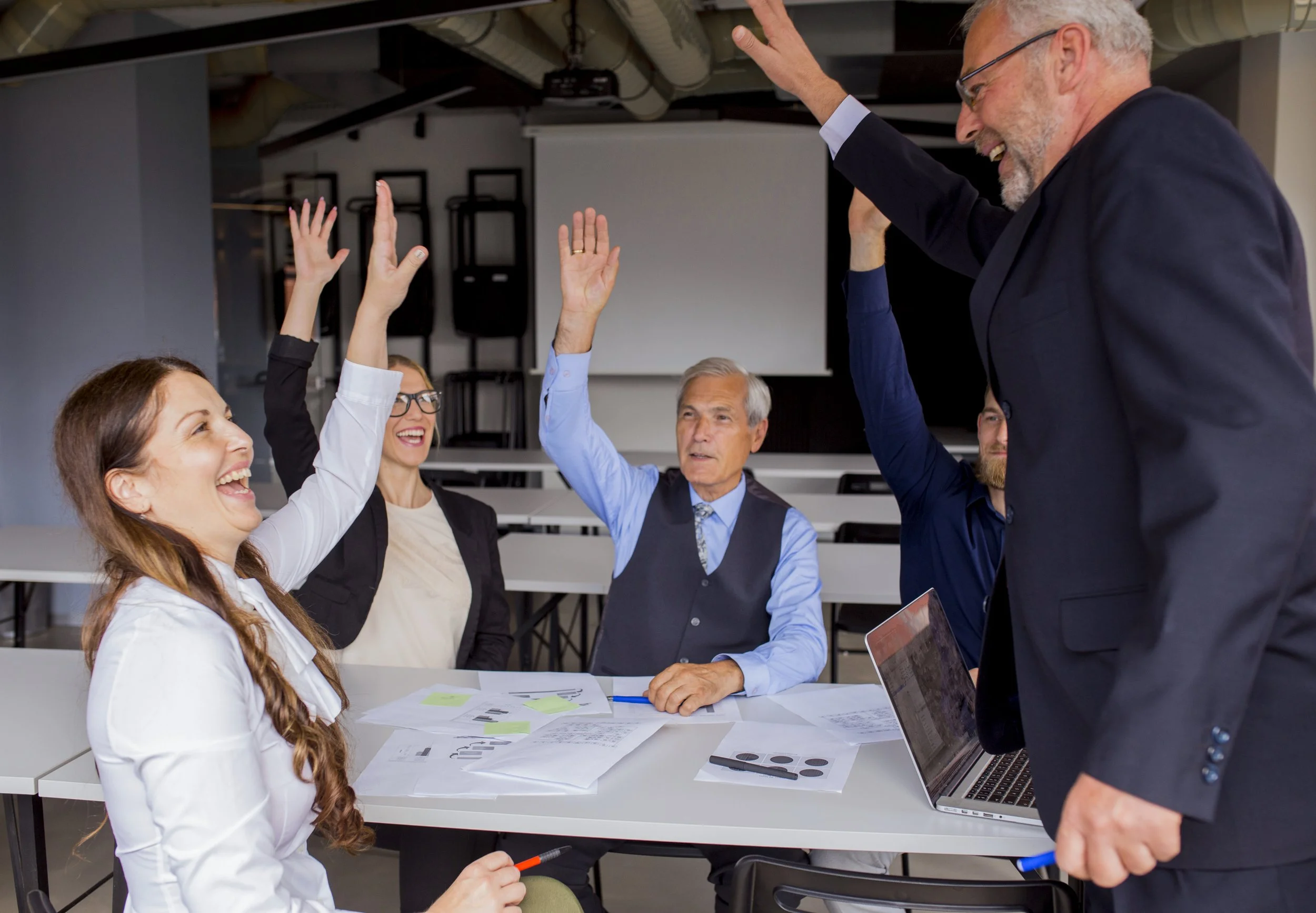 A diverse group of people in a meeting room raising their hands, with a man standing and smiling, possibly participating in a discussion or vote.