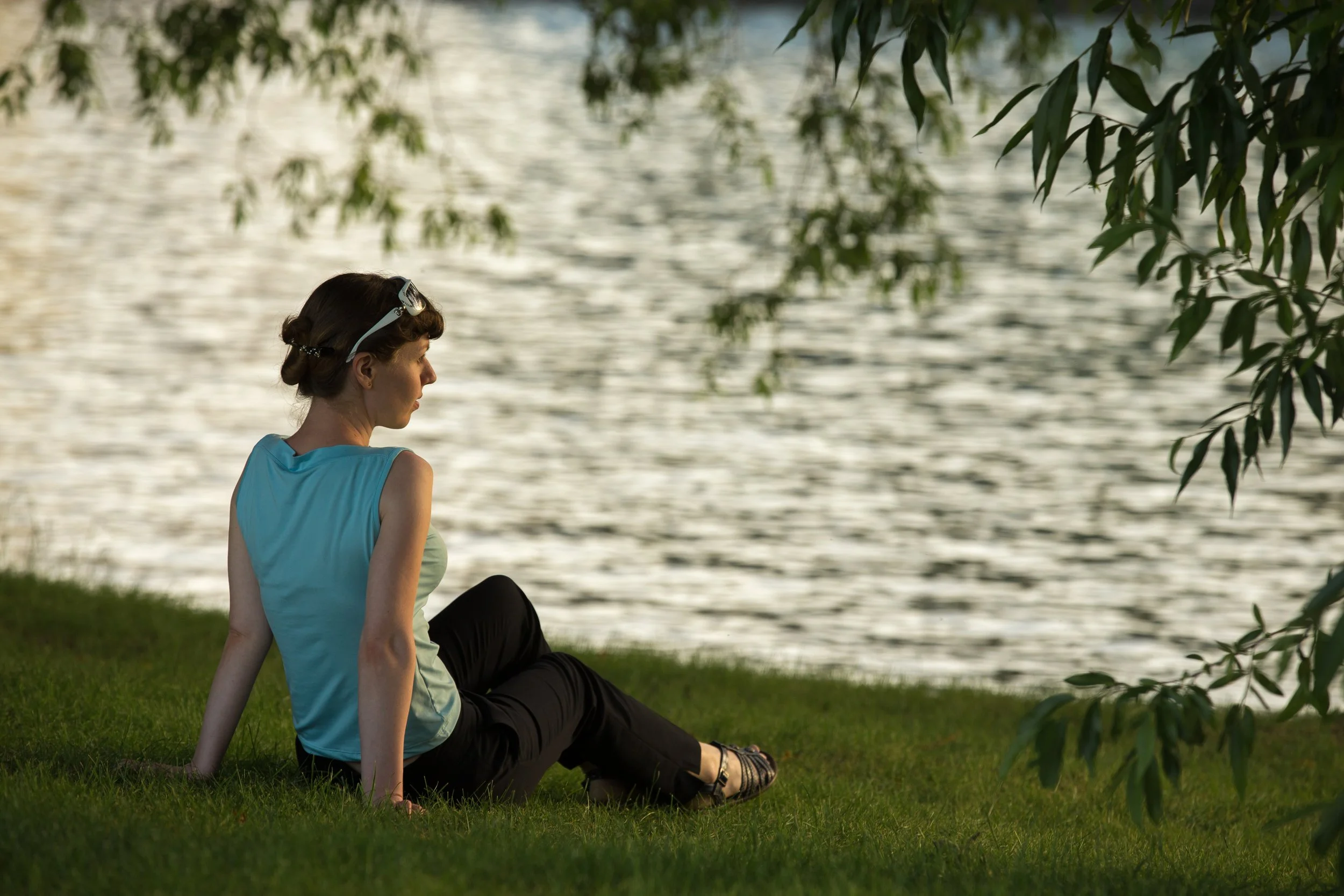 A woman sitting on grass near a body of water, with trees and leaves overhead, during what appears to be late afternoon or early evening.