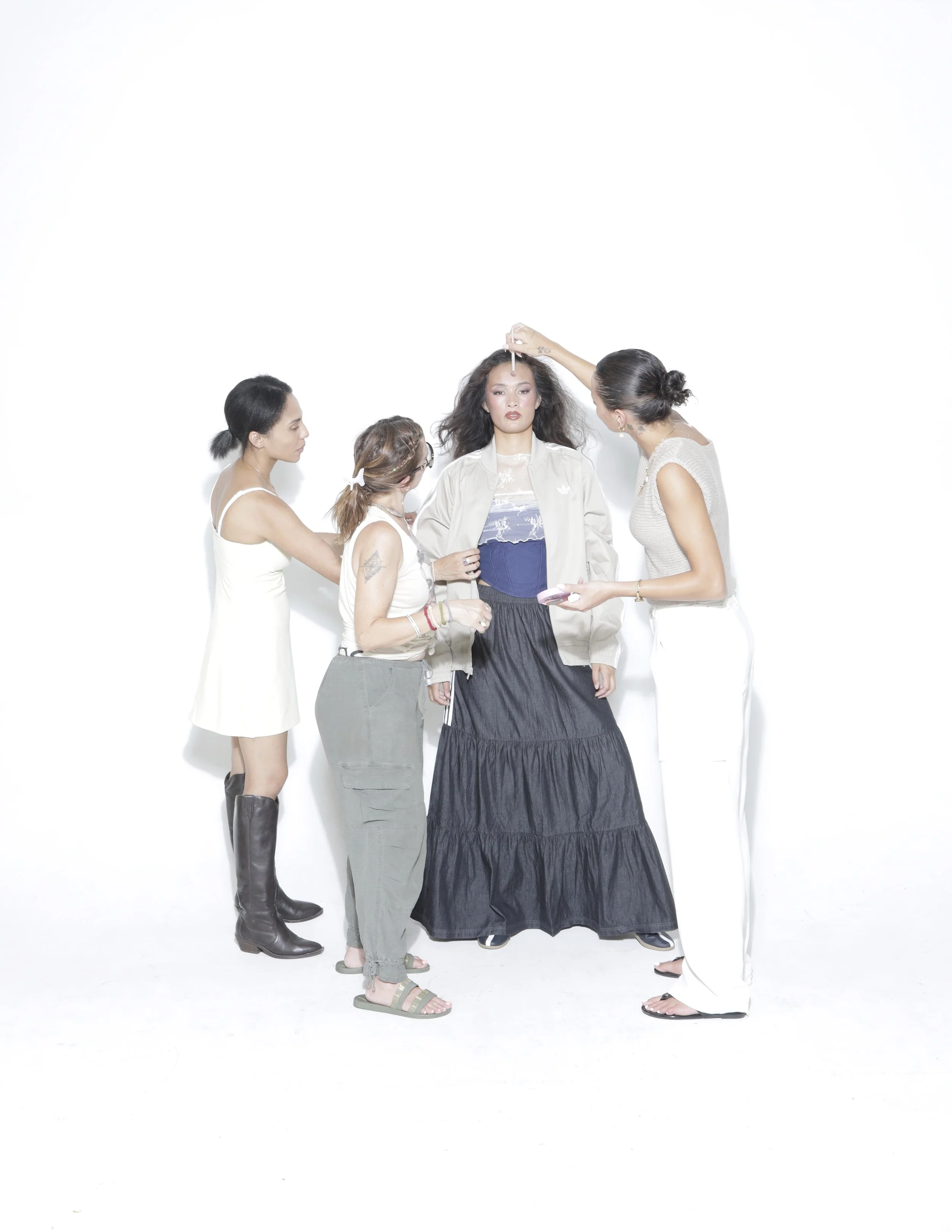 A woman with long dark hair standing with four women and a person, all with varying hairstyles, skin tones, and outfits, against a plain white background. The woman with long hair is being attended to with makeup or skincare products, while others assist or observe.