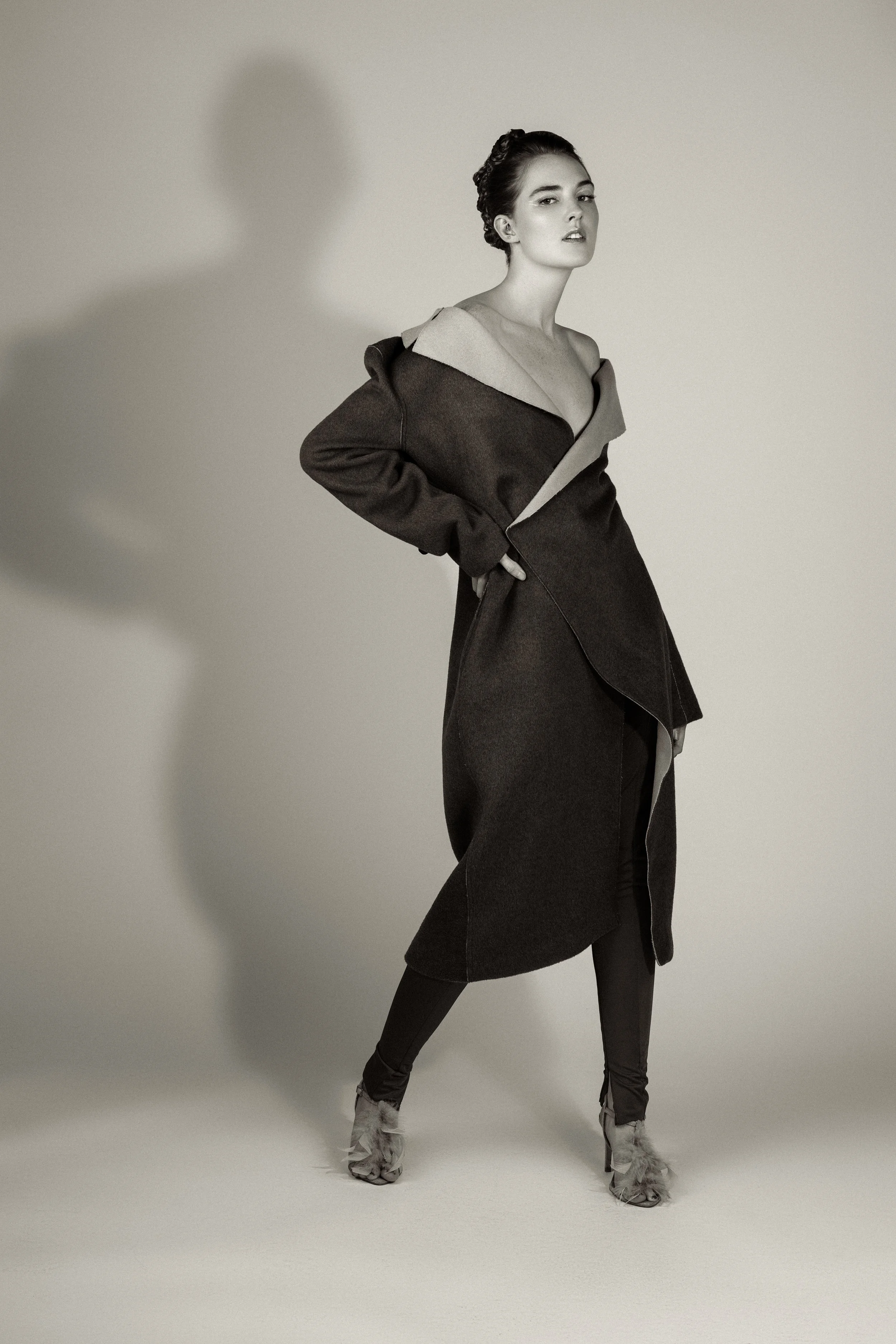A black and white photo of a woman posing in a studio with a plain background. She is wearing a dark, asymmetrical coat with a large collar, tights, and high-heeled shoes with feather details.