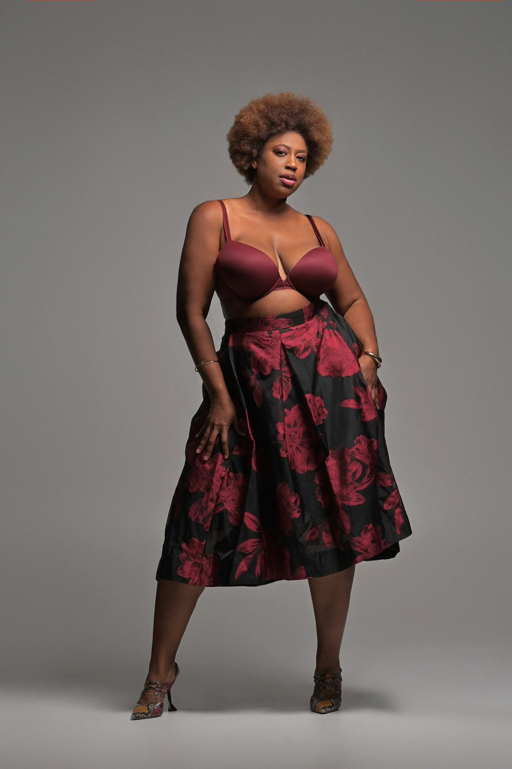 A confident woman with curly, natural hair stands against a plain gray background, wearing a burgundy bra, a floral-patterned skirt, and patterned high heels.