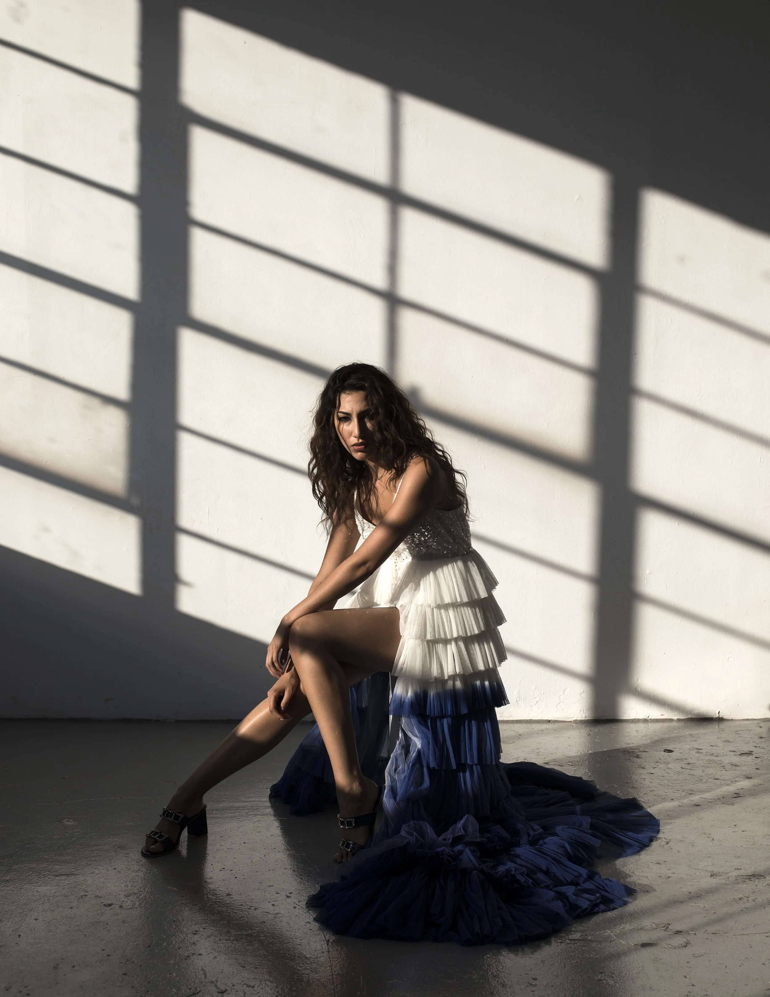 A woman with long curly hair wearing a layered white dress with a dark blue train, sitting on a chair in a room with a white wall and shadows from a window grid on the wall.