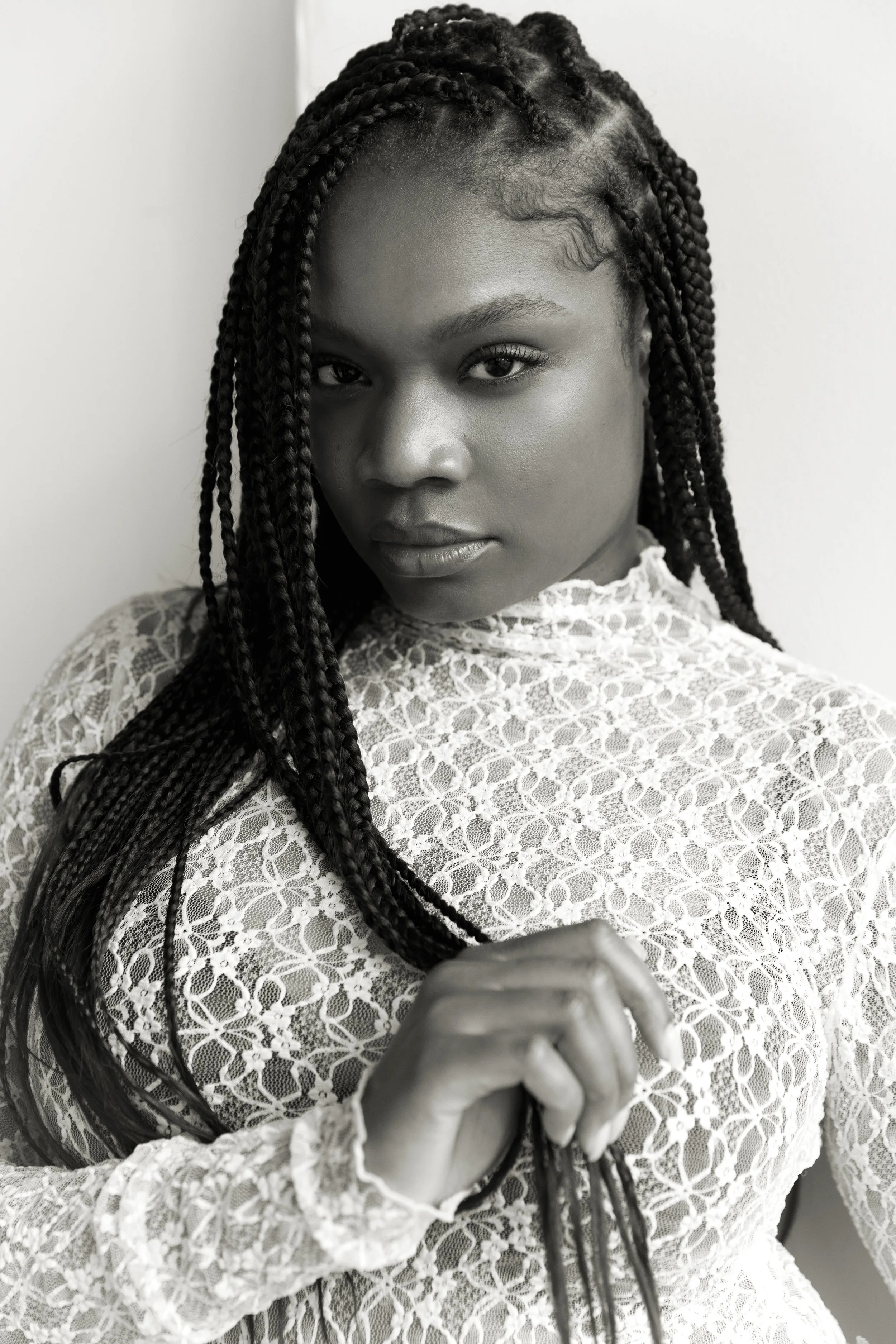 A woman with braided hair wearing a lace top, looking directly at the camera with a serious expression. Her hand is in the foreground, partially obscuring her face.