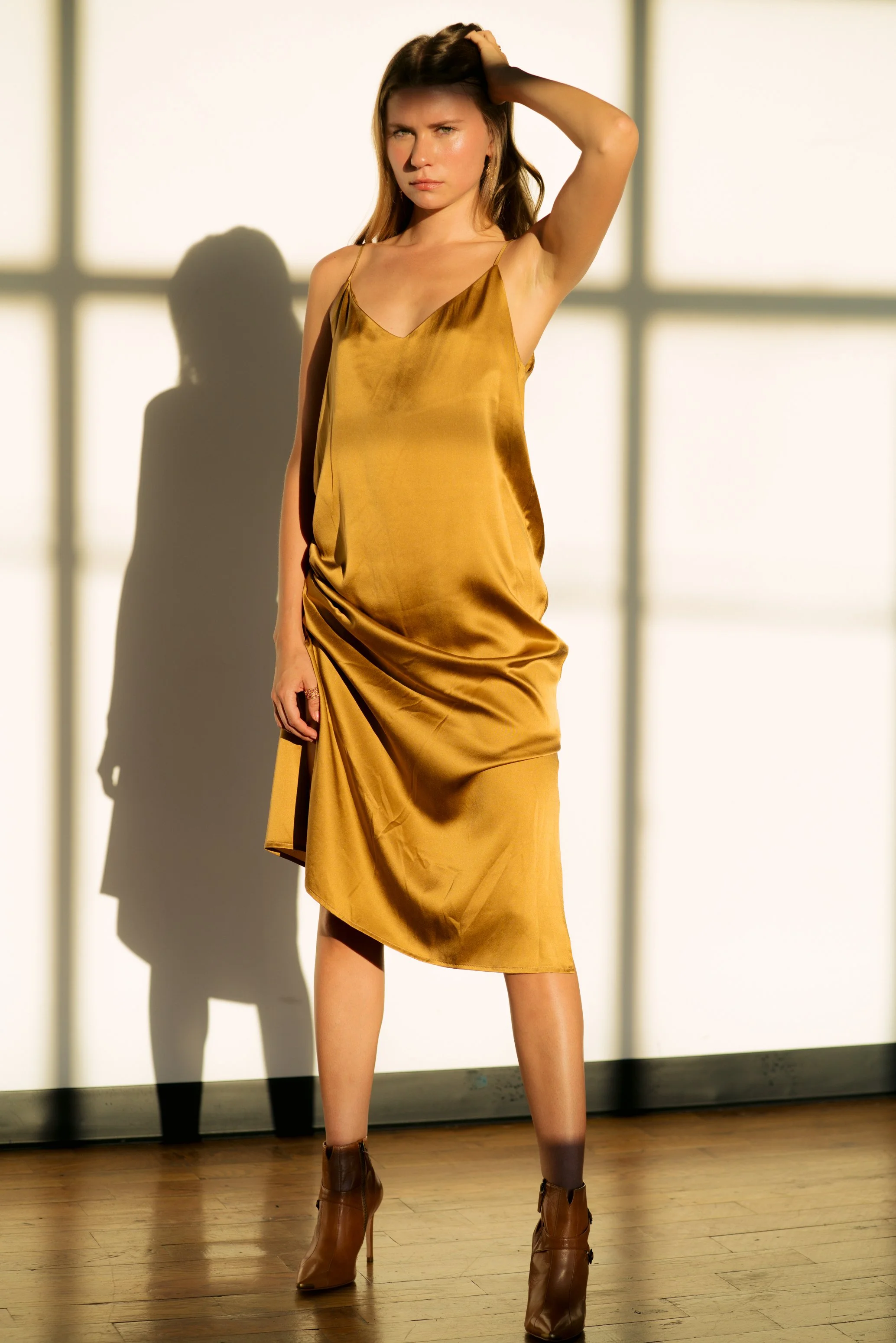 A woman standing on a wooden floor in front of a wall with large window shadows. She is wearing a gold-colored satin slip dress and brown high-heeled ankle boots. She has her left hand on her head, looking at the camera with a serious expression.