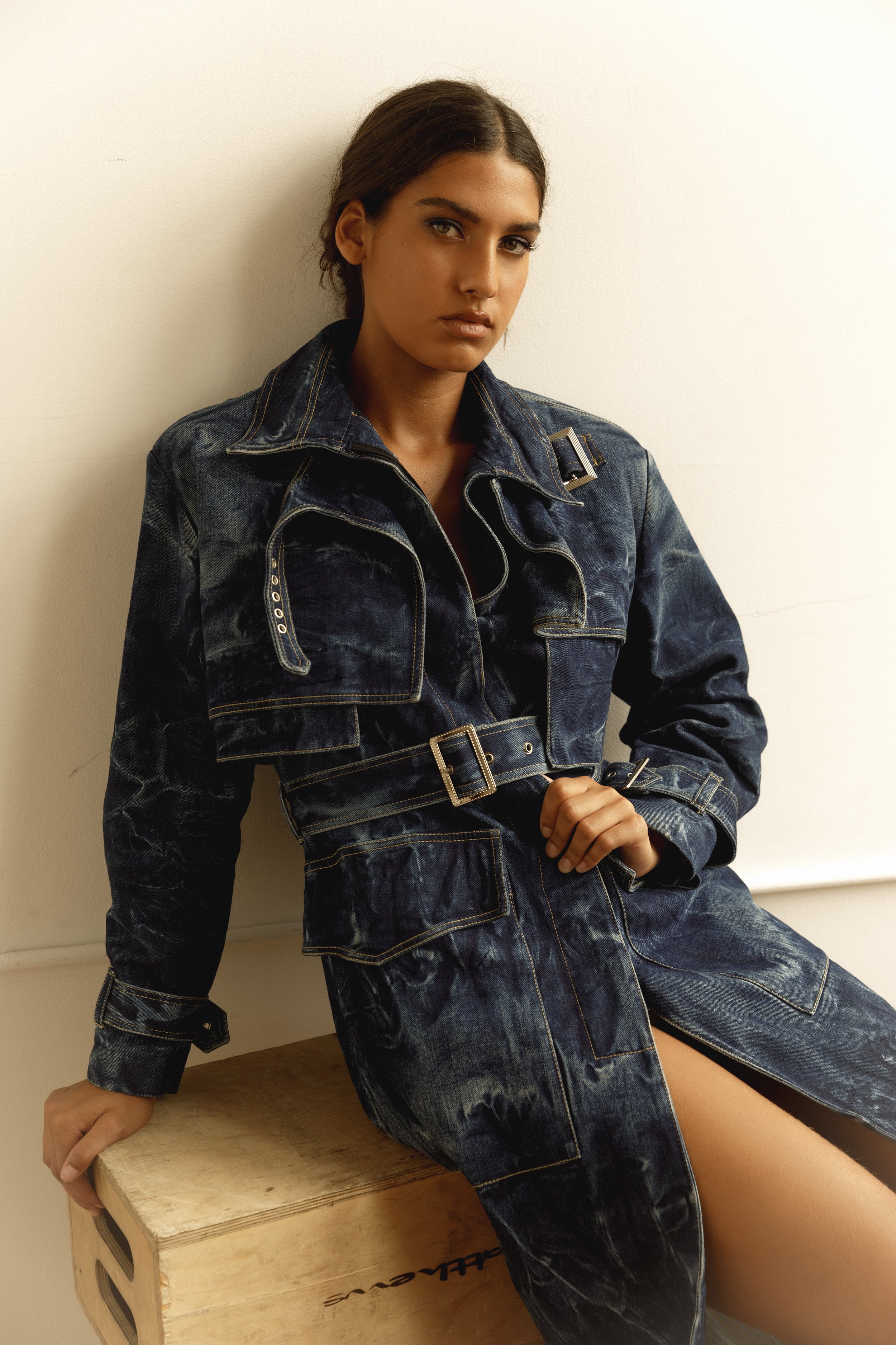 Woman with dark hair in a ponytail wearing a denim jacket and jeans, sitting on a wooden box against a plain white wall.