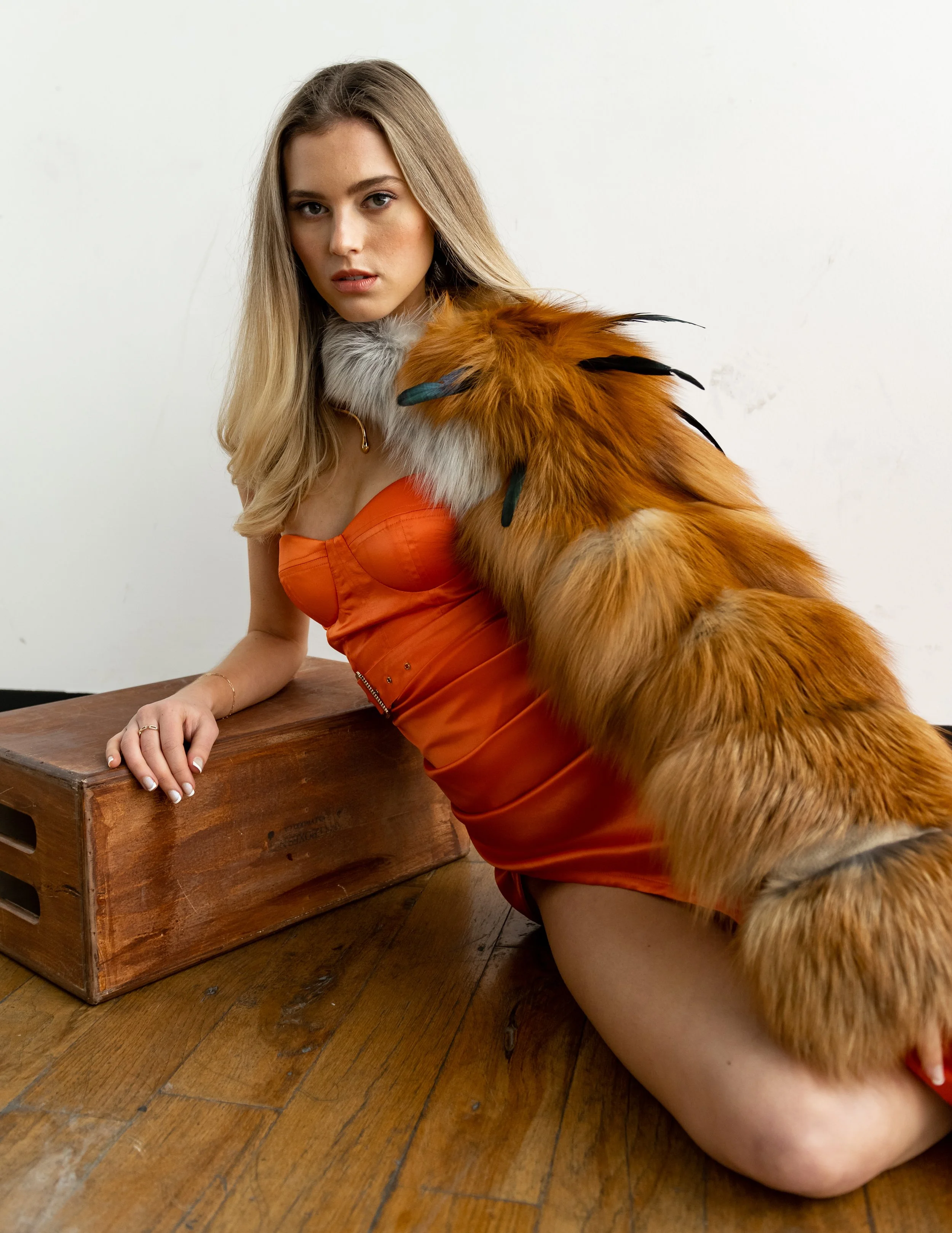A young woman with long blonde hair, wearing an orange dress and a faux fur stole with feathers, poses against a plain white background, kneeling on a wooden floor with one hand resting on a wooden box.