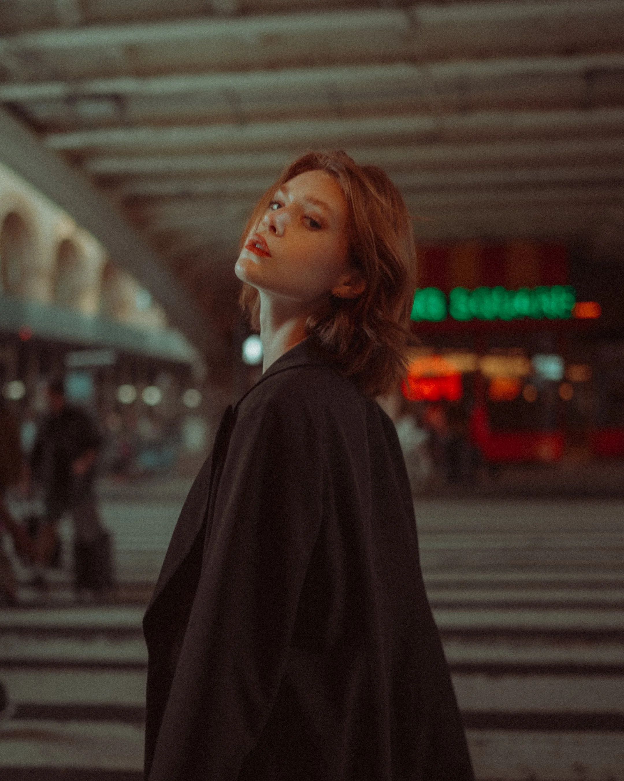 A woman with red hair and red lipstick stands in a parking garage with concrete ceiling and painted parking lines, looking over her shoulder.