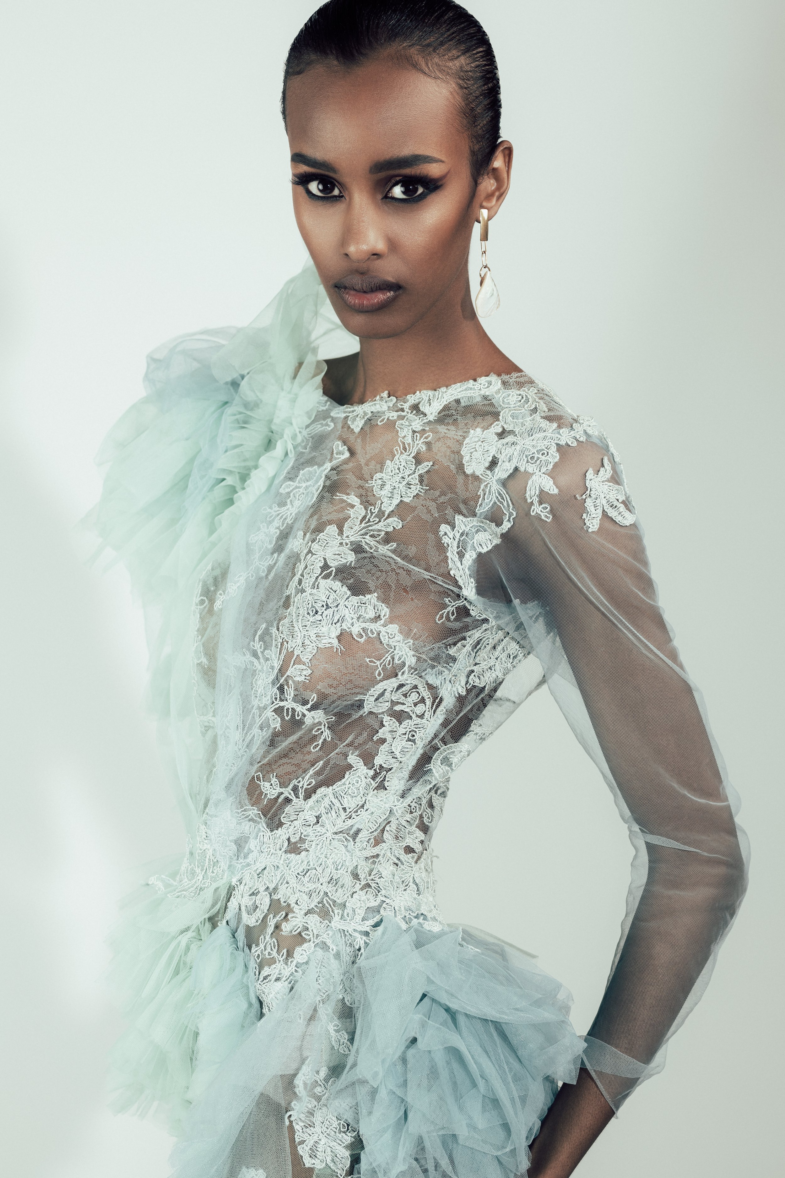 Woman with dark skin and slicked-back hair wearing a sheer, white, lace dress with ruffled and embroidered details, and a large white flower appliqué on her shoulder, posed against a plain background.