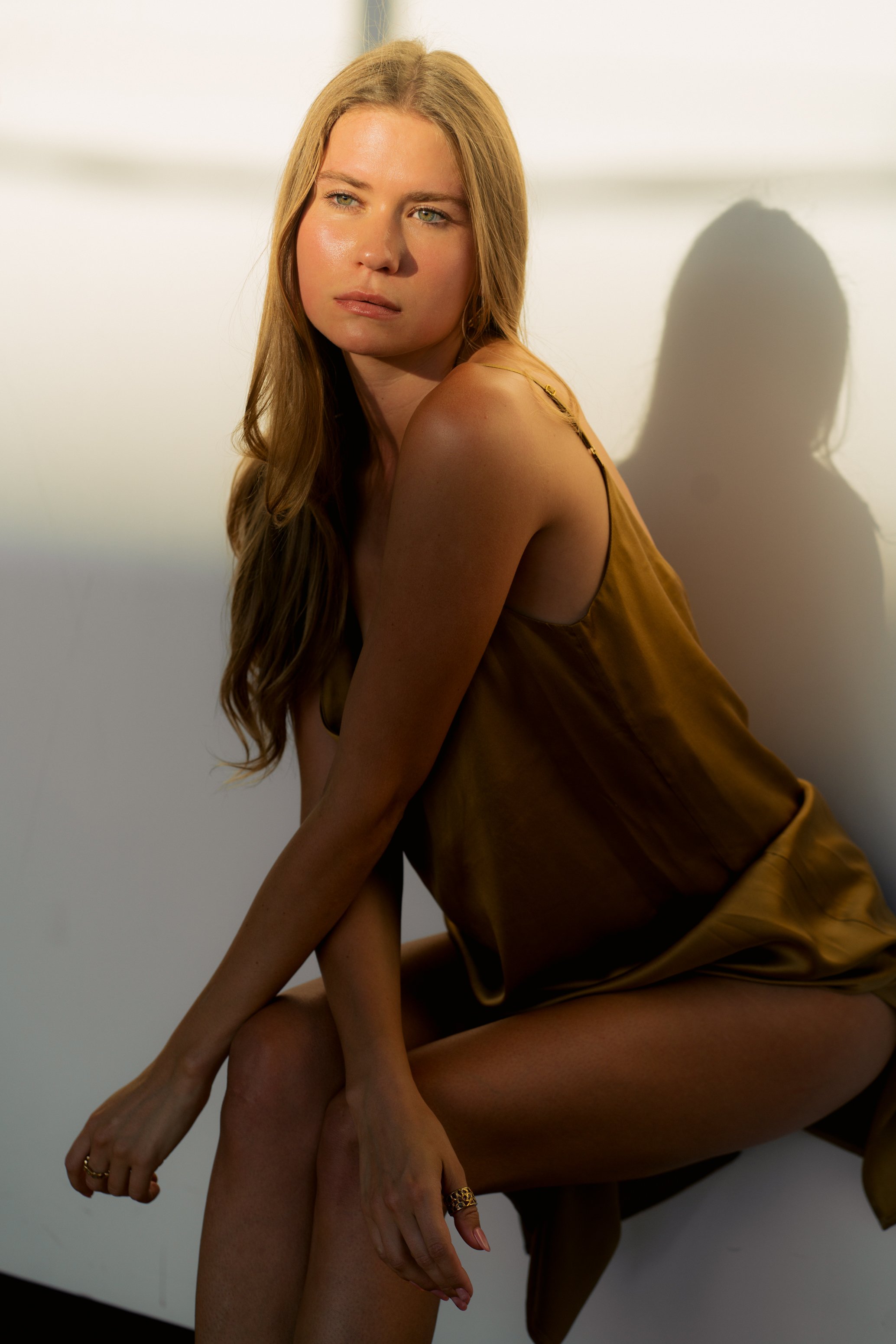 A woman with long blonde hair and blue eyes dressed in a silky, brownish-golden dress sitting against a white wall with a shadow cast on it.