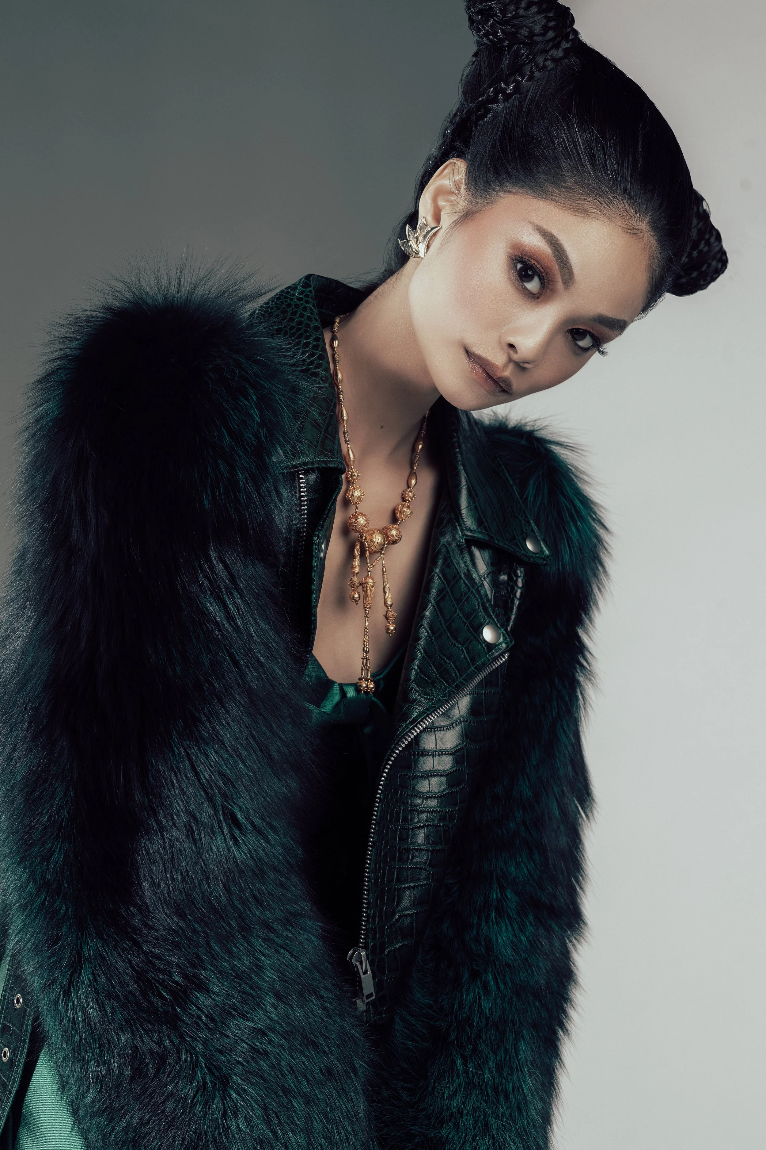 A woman with dark braided hair wearing a black leather jacket with fur trim, gold jewelry, and silver leaf-shaped earrings, posing against a plain background.