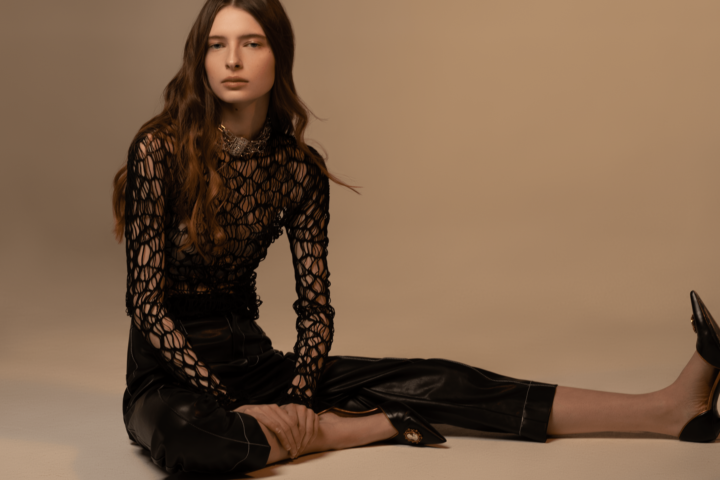 A woman with long brown hair, wearing a black open-knit top and black leather pants, is sitting on the floor against a beige background. She is wearing a sparkly necklace and black heeled shoes, with one leg extended and the other bent, resting her h