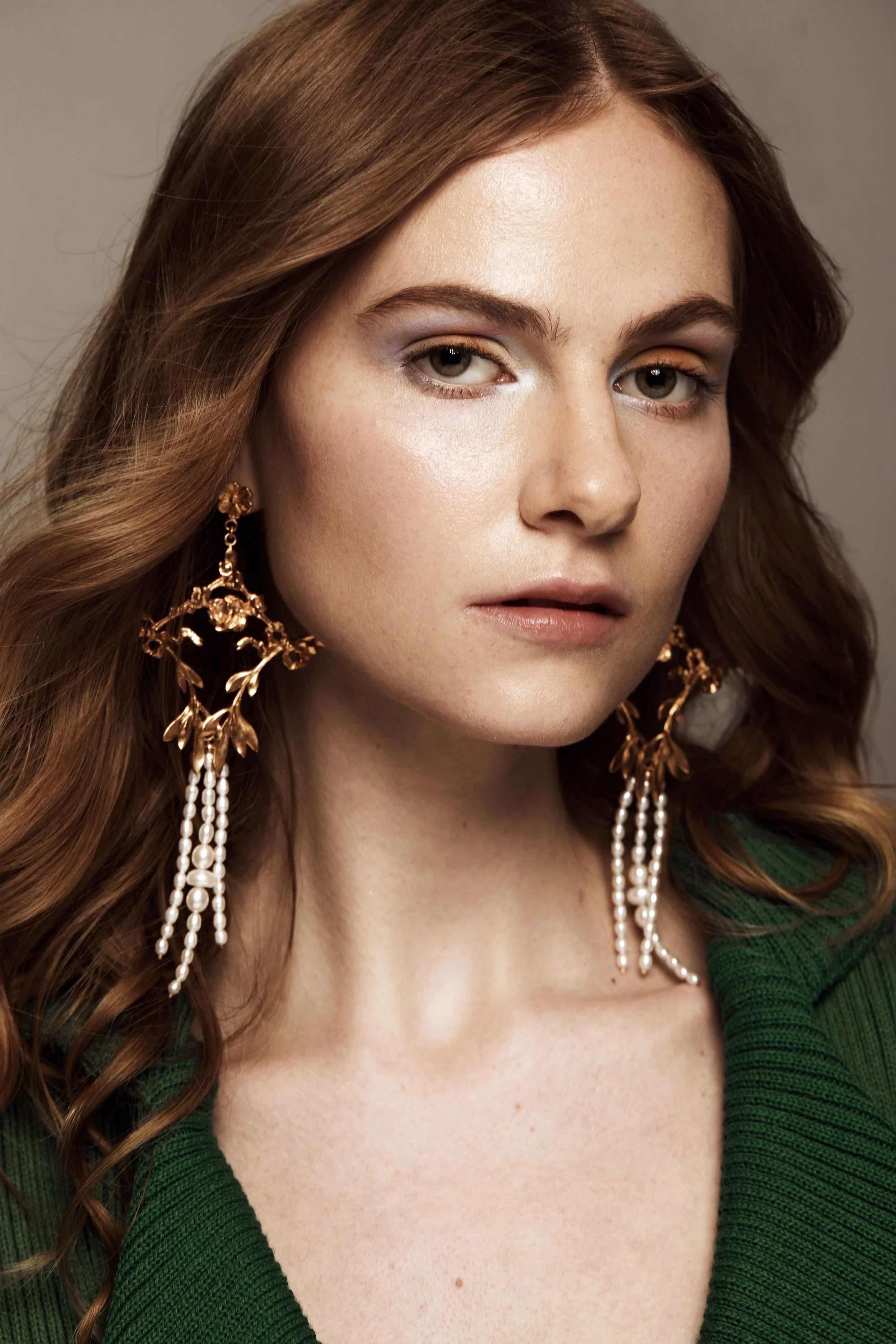 Close-up of a woman with red hair wearing makeup and large gold leaf earrings with pearl accents. She is dressed in a green garment.