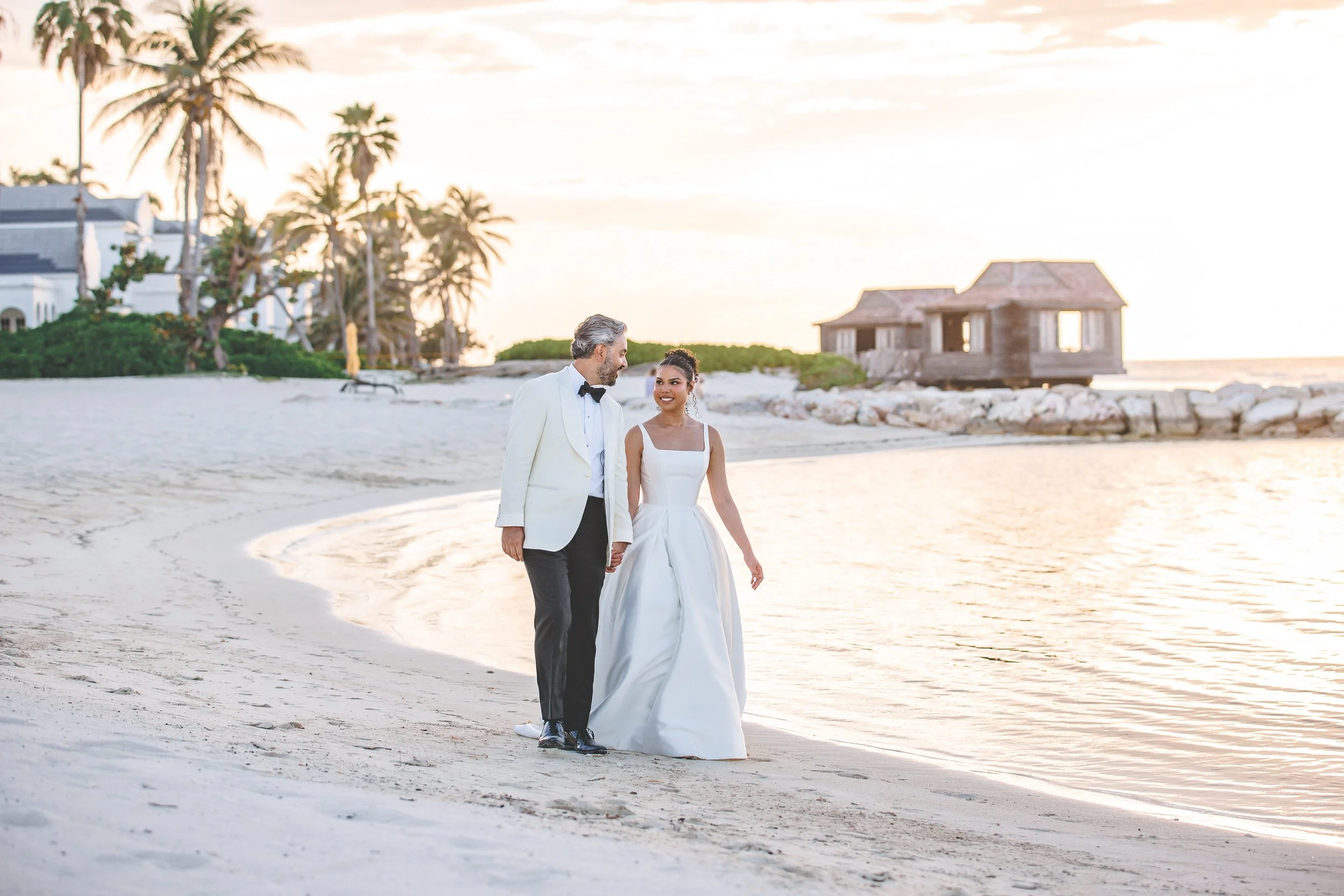 A groom in an ivory bespoke dinner jacket walks the beach at sunset with his bride