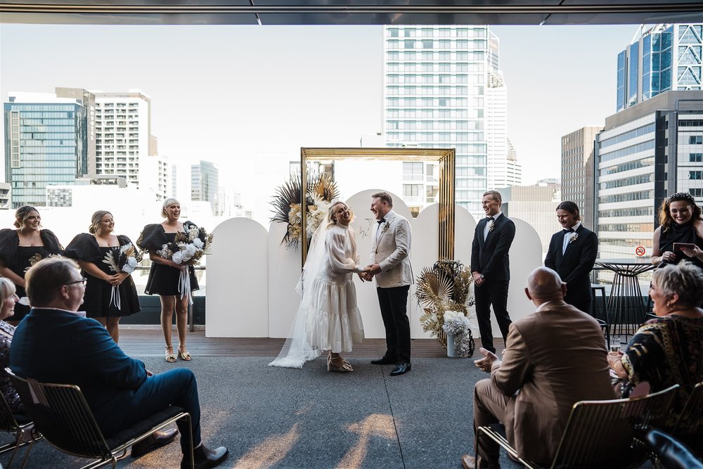  the couple saying their vows at each other in front of their guests in Aurora Rooftop Melbourne Hotel 
